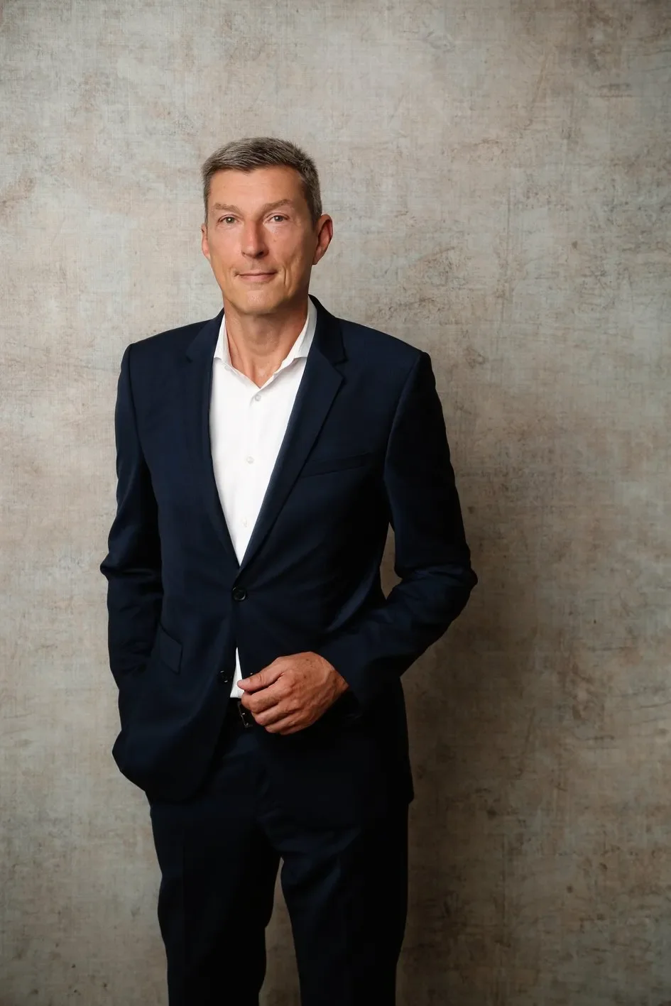 Ein Mann in einem schwarzen Anzug und weißem Hemd steht vor einer hellbraunen Wand. Er hat kurze, graue Haare und schaut ernst in die Kamera.