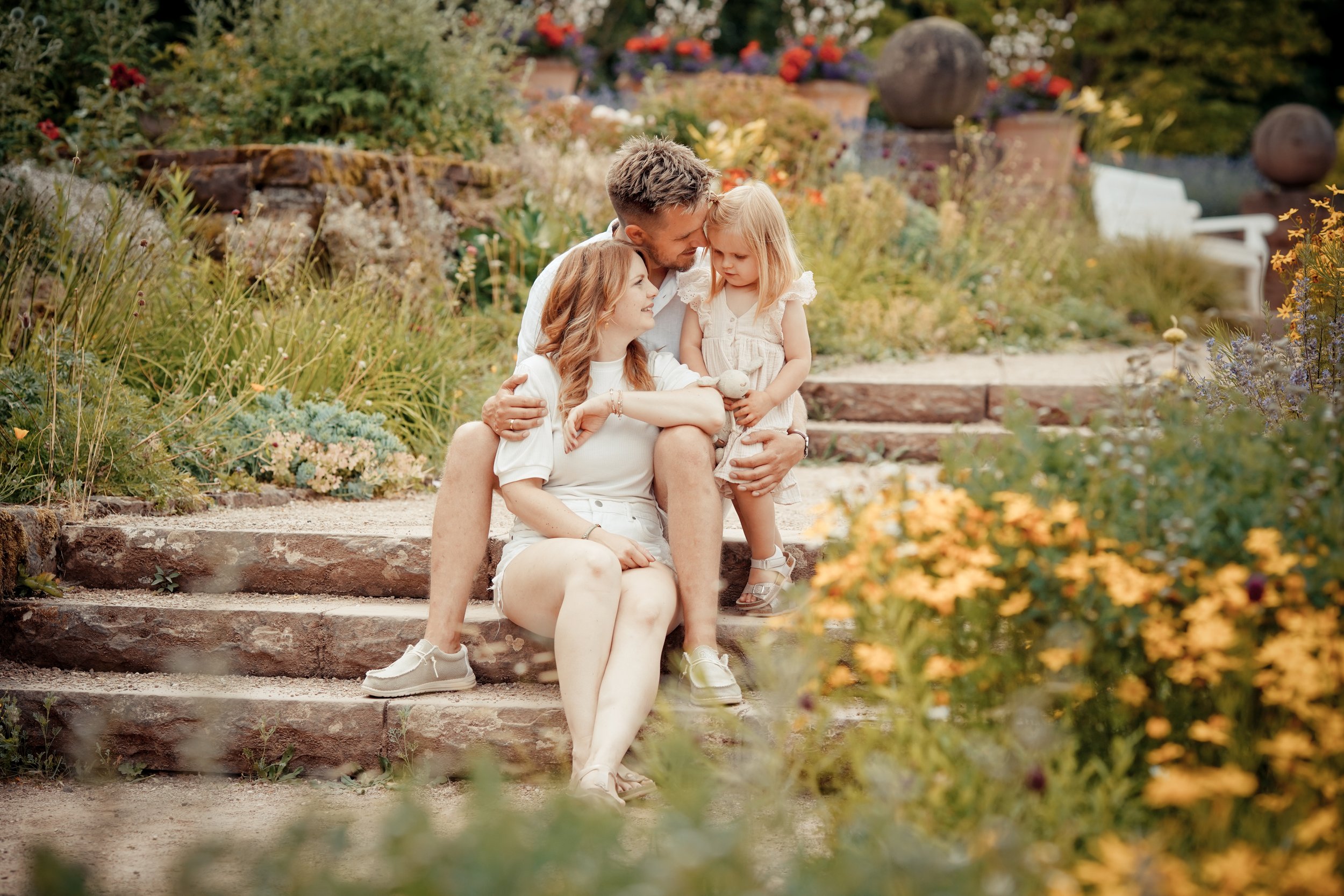 Familie mit zwei Erwachsenen und einem Kind sitzt auf Steinen im Garten, umgeben von Pflanzen und Blumen.