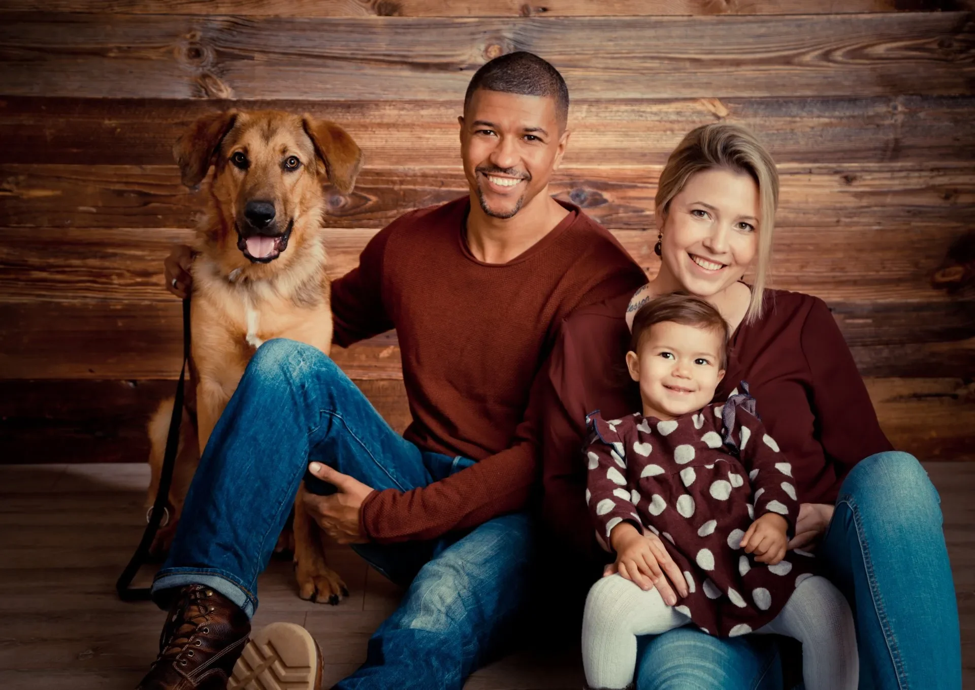Eine glückliche Familie mit einem Hund, bestehend aus einem Mann, einer Frau und einem Kind, sitzen vor einer Holzverkleidung. Alle lächeln freundlich in die Kamera.