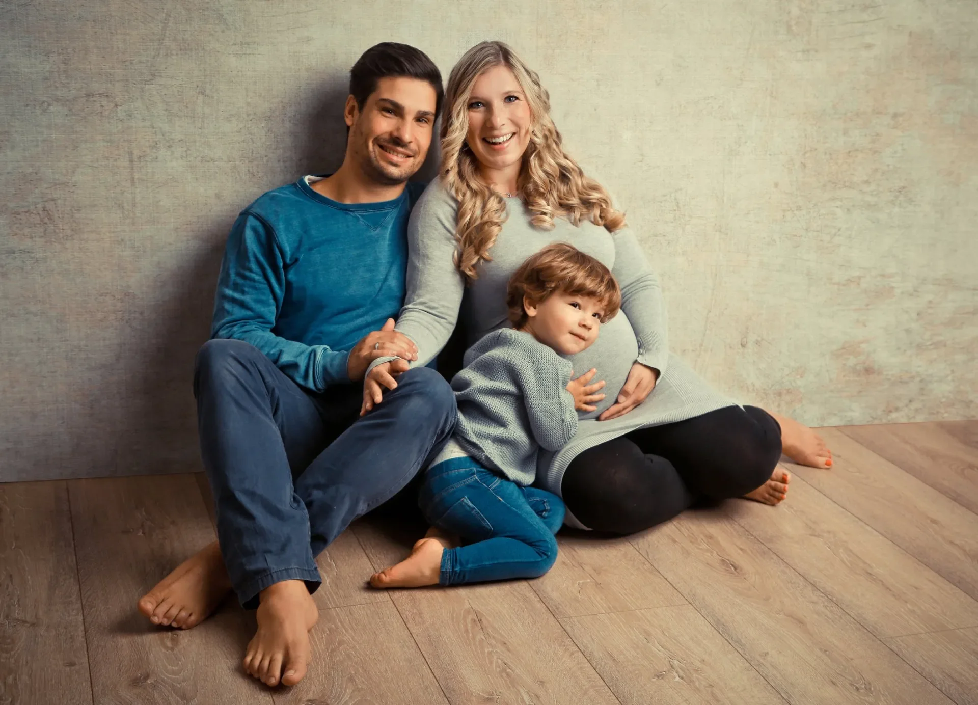 Familienfotoshooting im Studio. Eine glückliche Familie sitzt und liegt lächelnd auf einem Holzboden vor einer neutralen Wand. Der Vater, die Mutter und ein kleines Kind umarmen sich. 