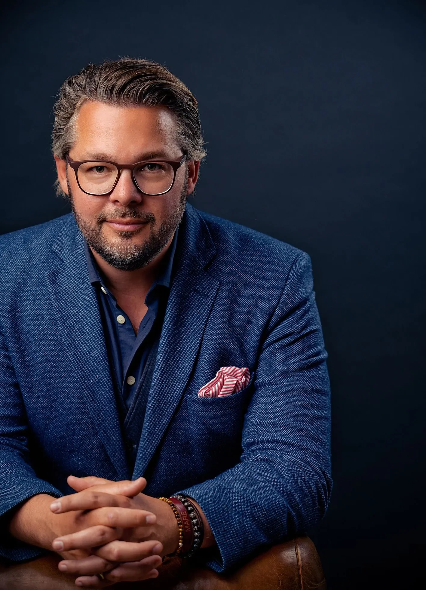 Ein Mann mit Brille, grauem Haarschopf und Bart trägt einen blauen Anzug und sitzt vor einem dunklen Hintergrund.