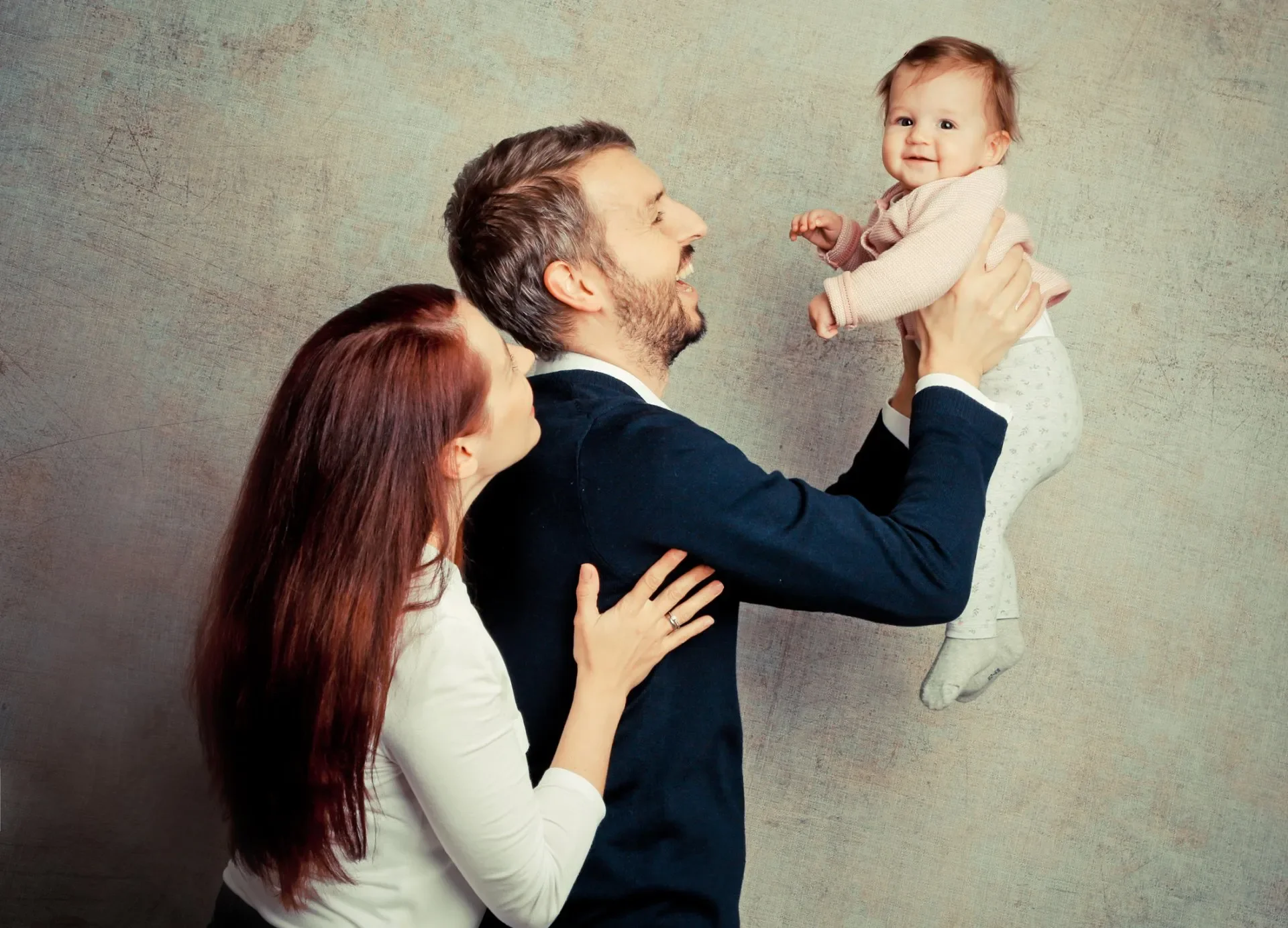 Familien Portrait Fotoshooting Familie besteht aus Vater, Mutter und Baby, sie lachen und genießen gemeinsame Zeit, Vater hebt das Baby hoch, Mutter steht neben ihnen, im Hintergrund eine Wand mit Textur.