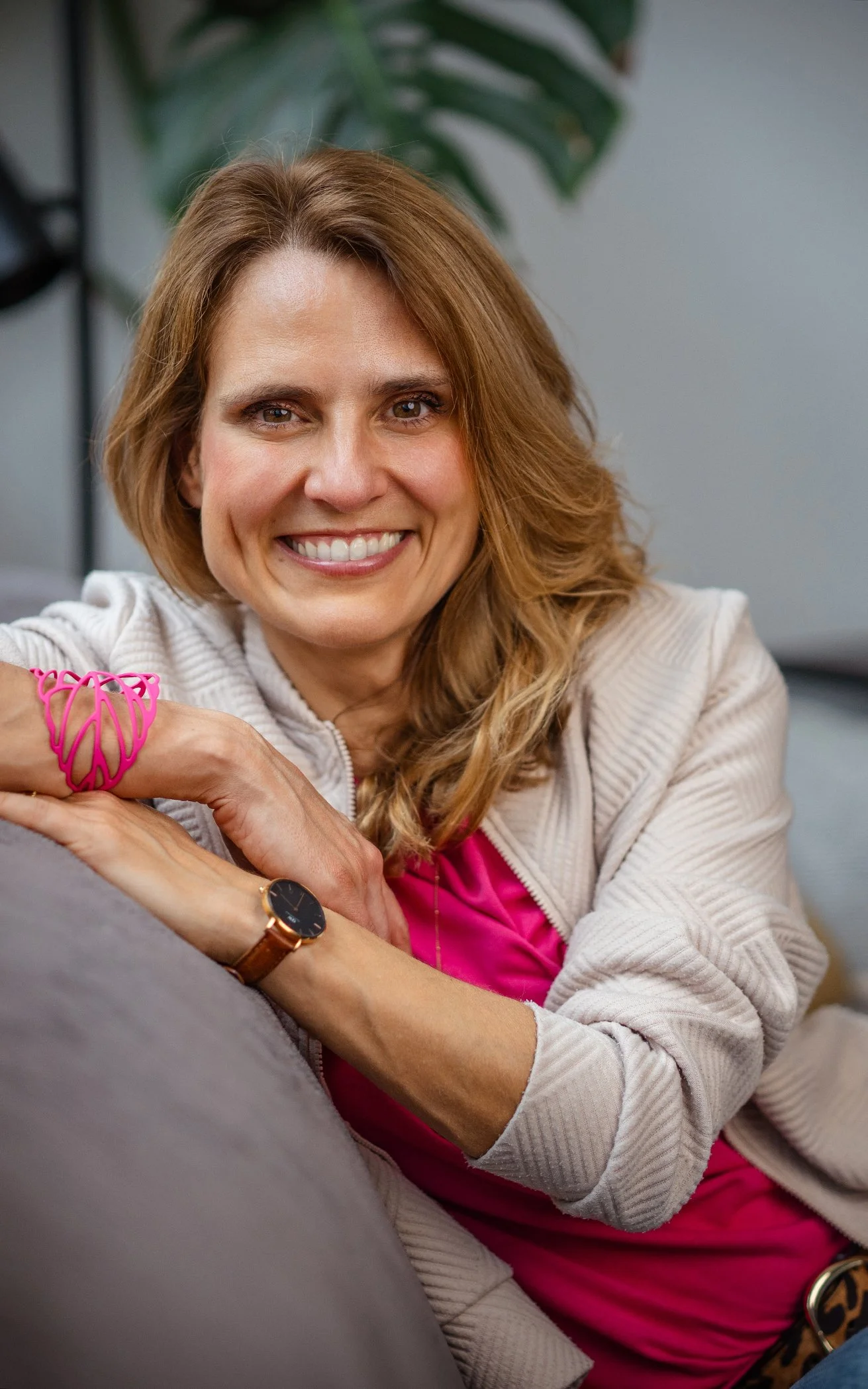 Lächelnde Frau mit lockigem, rotbraunem Haar sitzt auf einer Couch und schaut erfreut in die Kamera, trägt ein pinkes Hemd, ein beiges Jackett, eine Armbanduhr und eine pinke Schmuckschlange am Handgelenk, im Hintergrund eine große Pflanze.