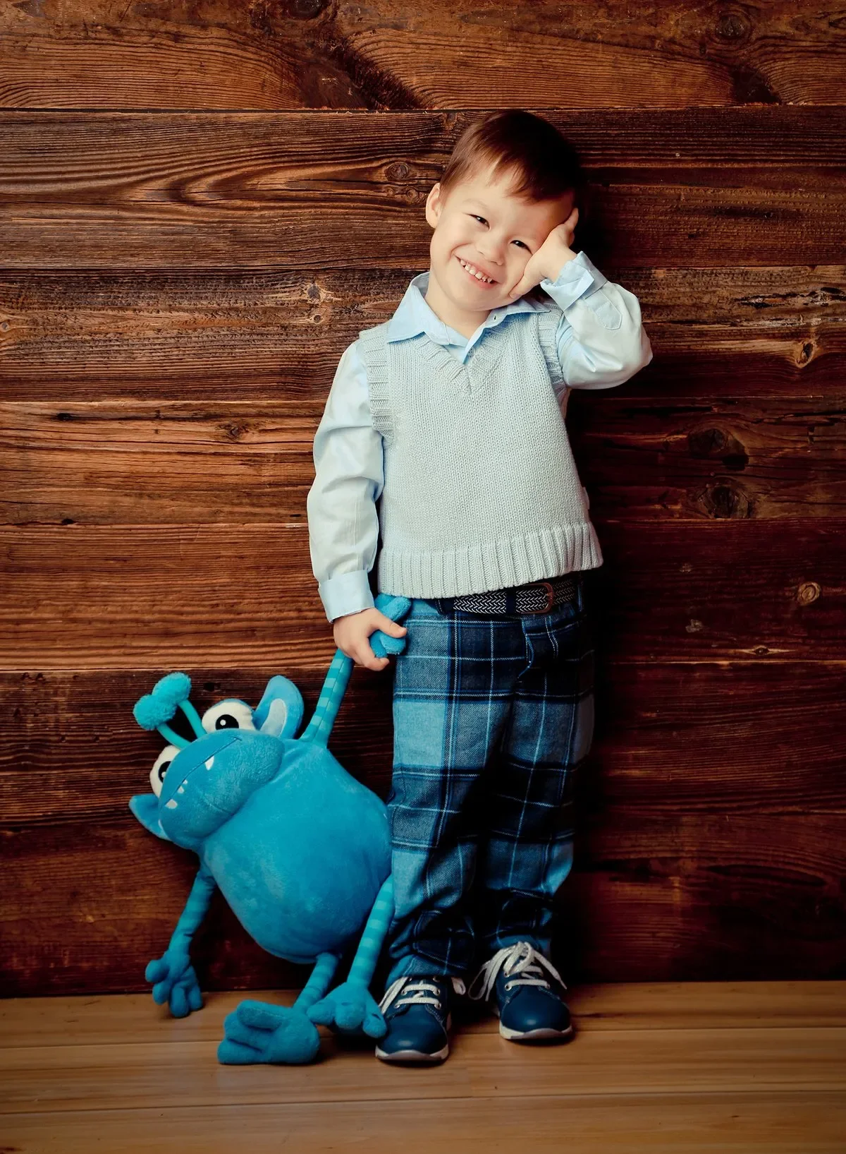 Kind beim Fotoshooting - Ein lachender Junge im Hemd, Sweater, Jeans und Turnschuhen steht vor einer Holzwand. Er hält eine blaue Monster-Tasche mit Armen und Beinen. 