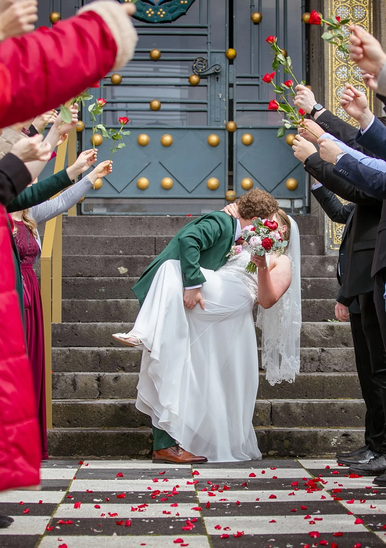 Hochzeitsszene vor einer dicken Treppe mit Rosenblättern auf dem Boden. Ein Brautpaar, der Bräutigam in grünem Anzug und die Braut in weißem Kleid, küsst sich. Freunde und Familie grüßen mit Rosen.