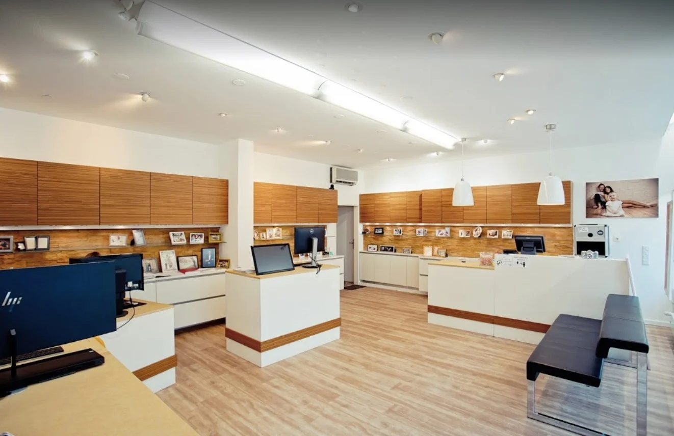 Räumlichkeiten von Fotostudio Hirch eines modernen Studios mit weißen Wänden, Holzlaminatboden, mehreren Empfangstresen mit Computern, Bilderrahmen und Dekorationen, Sitzbank aus schwarzem Leder, Wandbild eines Paares, Deckenleuchten und Klimaanlage.