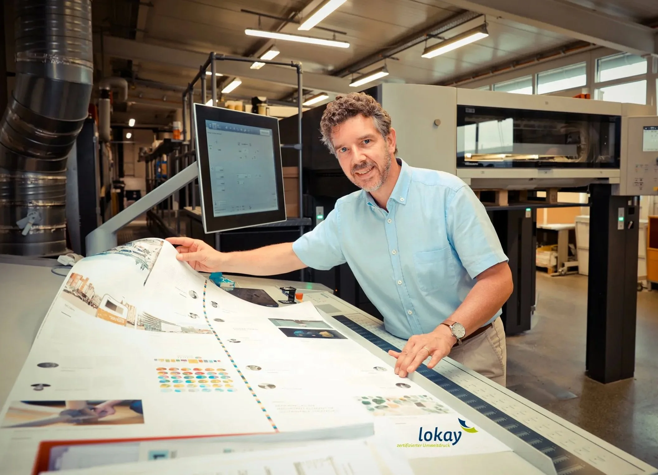 Mann schaut in ein großes aufgeschlagenes Poster mit Farbdarstellungen und Text, in einer Druckerei oder Labor. Im Hintergrund industrielle Geräte, Computerbildschirm und helle Fenster.