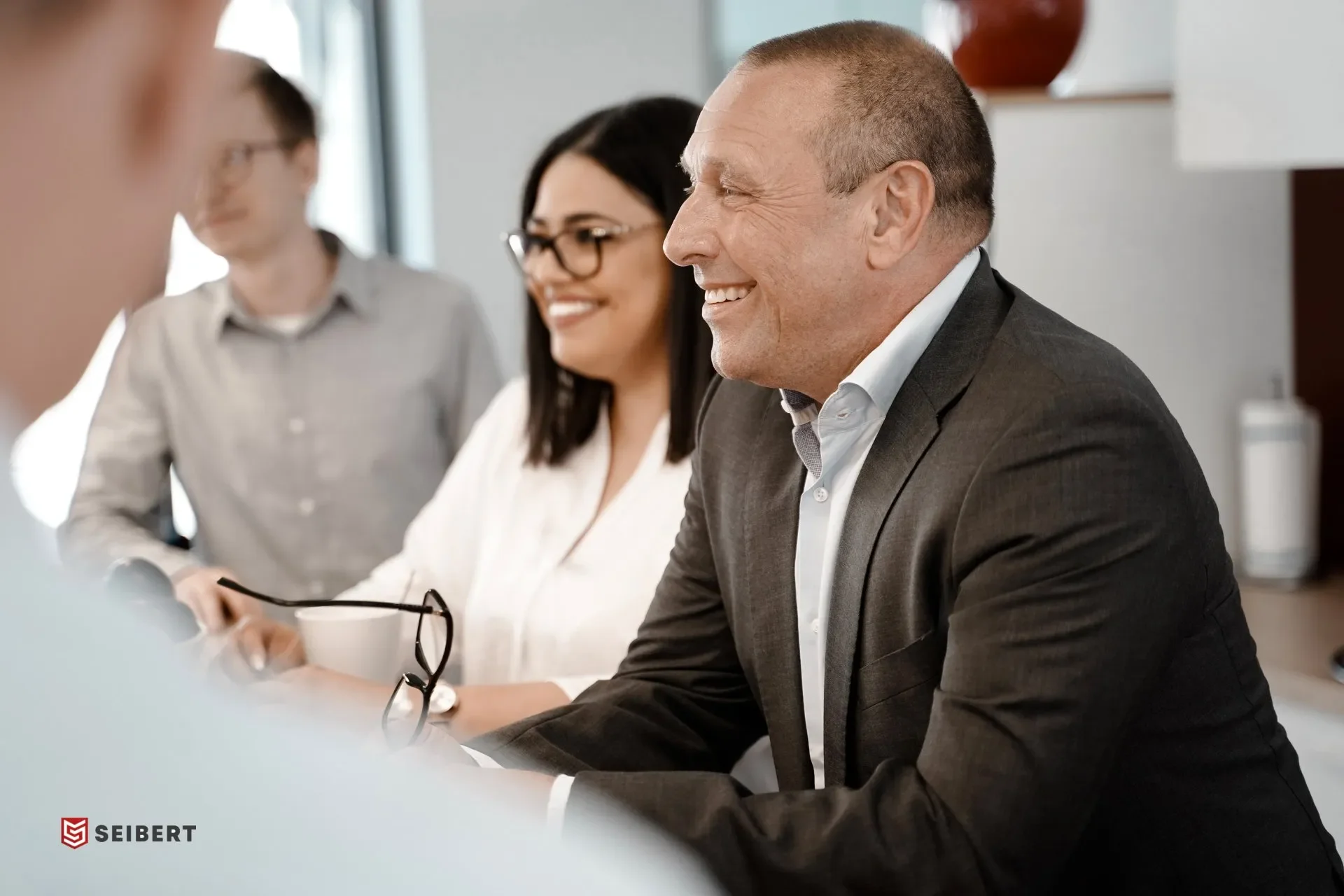 Firmenbild vor Ort im Büro. Menschen bei einem Meeting, frontal Blick auf einen glücklichen Mann im Anzug, im Hintergrund eine Frauen jüngere und ältere, die lachen und sich unterhalten.