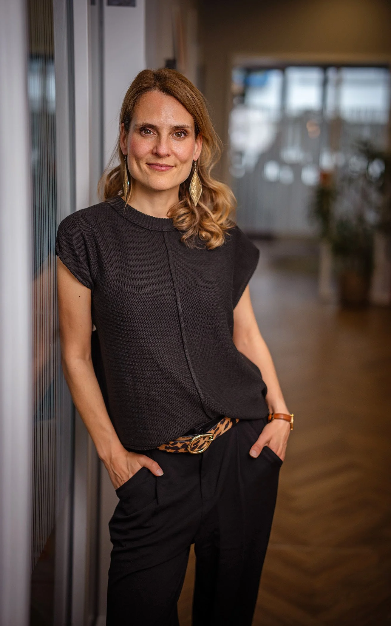 Business Portrait Eine Frau mit braunen, gewellten Haaren, die ein schwarzes Oberteil trägt und eine Leoparden-Gürtel trägt, lehnt an einer Wand in einem modernen Innenraum.