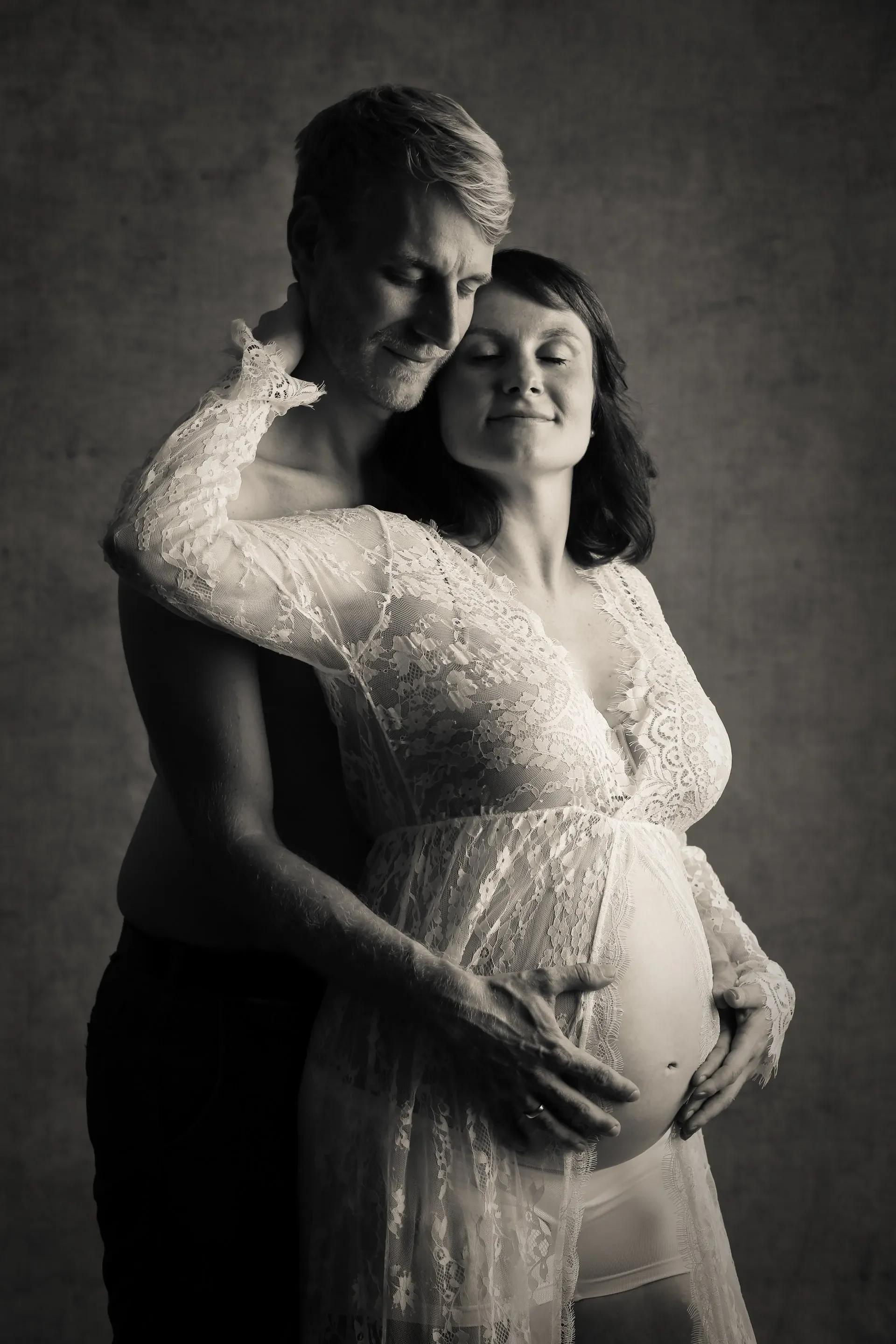 Babybauch Fotoshooting im Studio - Schwarz-Weiß-Foto eines schwangeren Frau mit einem Mann, beide mit geschlossenen Augen, in einem intimen Moment, die Frau trägt ein weißes Spitzenkleid, der Mann hält sie liebevoll umarmt.