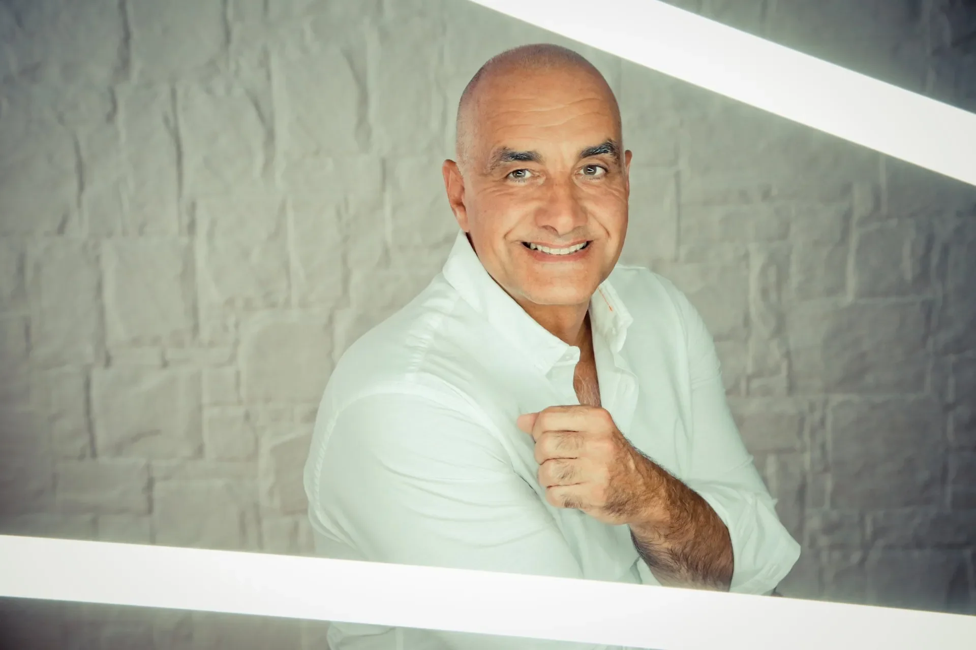 Business Portrait Mann mit weißem Hemd, lächelnd, schaut in die Kamera, vor einer Ziegelwand, scenisch beleuchtet.
