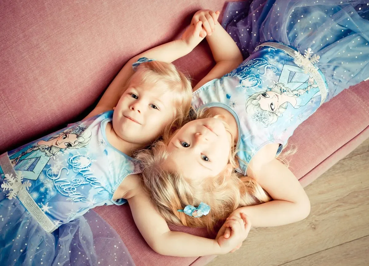 Zwei kleine Mädchen in Elsa-Kostümen, die auf einer Couch liegen und die Köpfe nebeneinander haben.