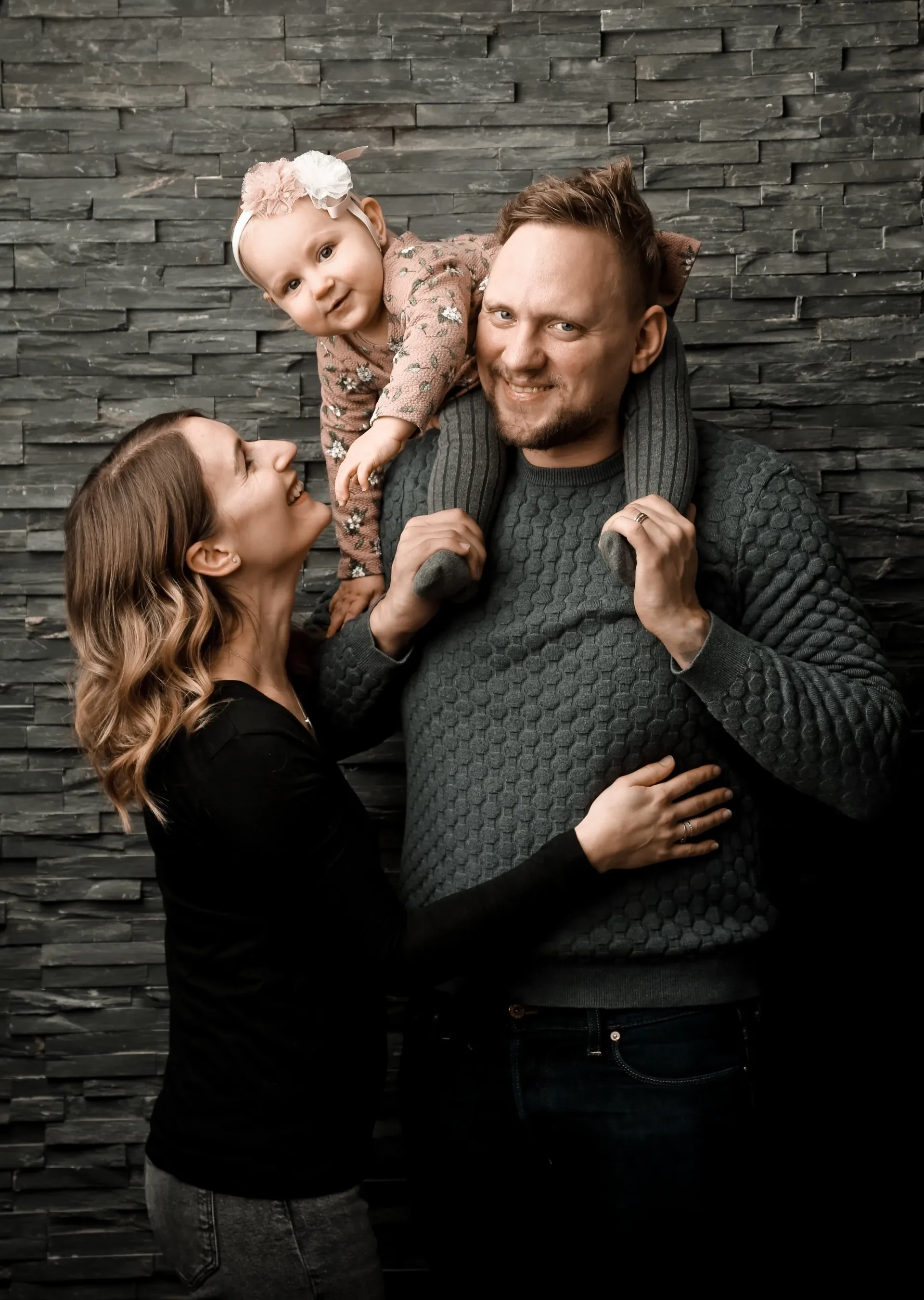 Familien Portrait Fotoshooting - Familie mit Tochter posiert vor einer Steinmauer, die Tochter sitzt auf den Schultern des Vaters, die Mutter küsst sie.