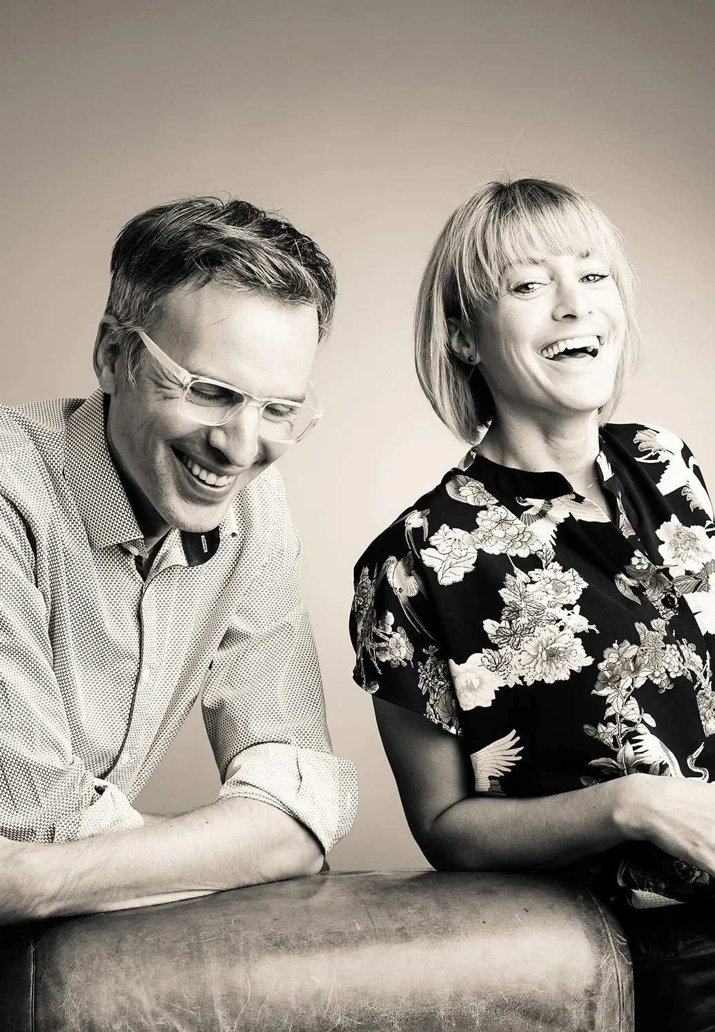 Geschäftsführer des Fotostudio Hirch. Zwei lachende Menschen, ein Mann mit Brille und ein elegantes Kleid, sitzen nebeneinander gegen einen neutralen Hintergrund. Sie lachen gemeinsam.