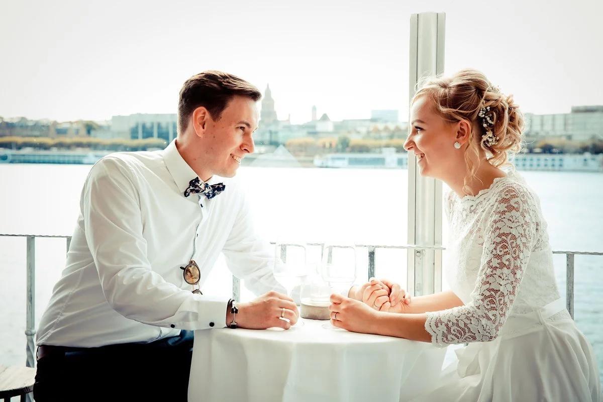 Hochzeitsfoto - Ein junges Paar sitzt gegenüber auf einer Terrasse bei einem romantischen Essen, hält Händchen und lächelt sich an. Im Hintergrund ist eine Stadtansicht mit Fluss und Gebäuden zu sehen.