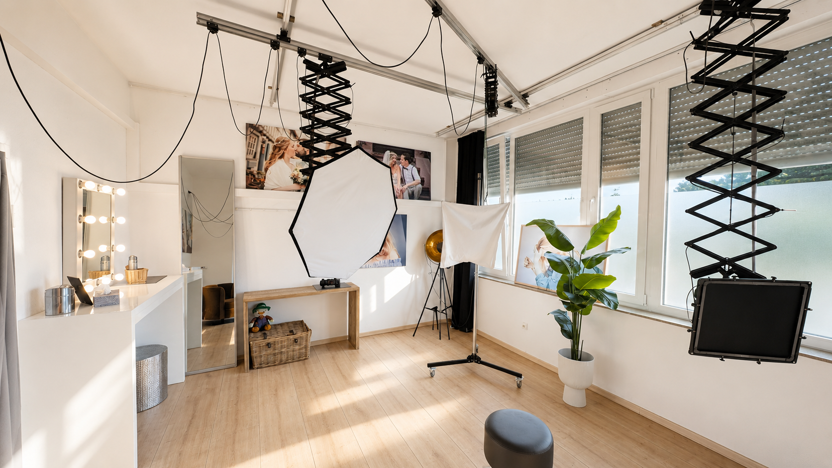 Innenraum eines Fotostudios mit Beleuchtungs- und Fotografieausrüstung, großem Fenster, Spiegel, Bilddekorationen an der Wand, Pflanze und Sitzhocker.
