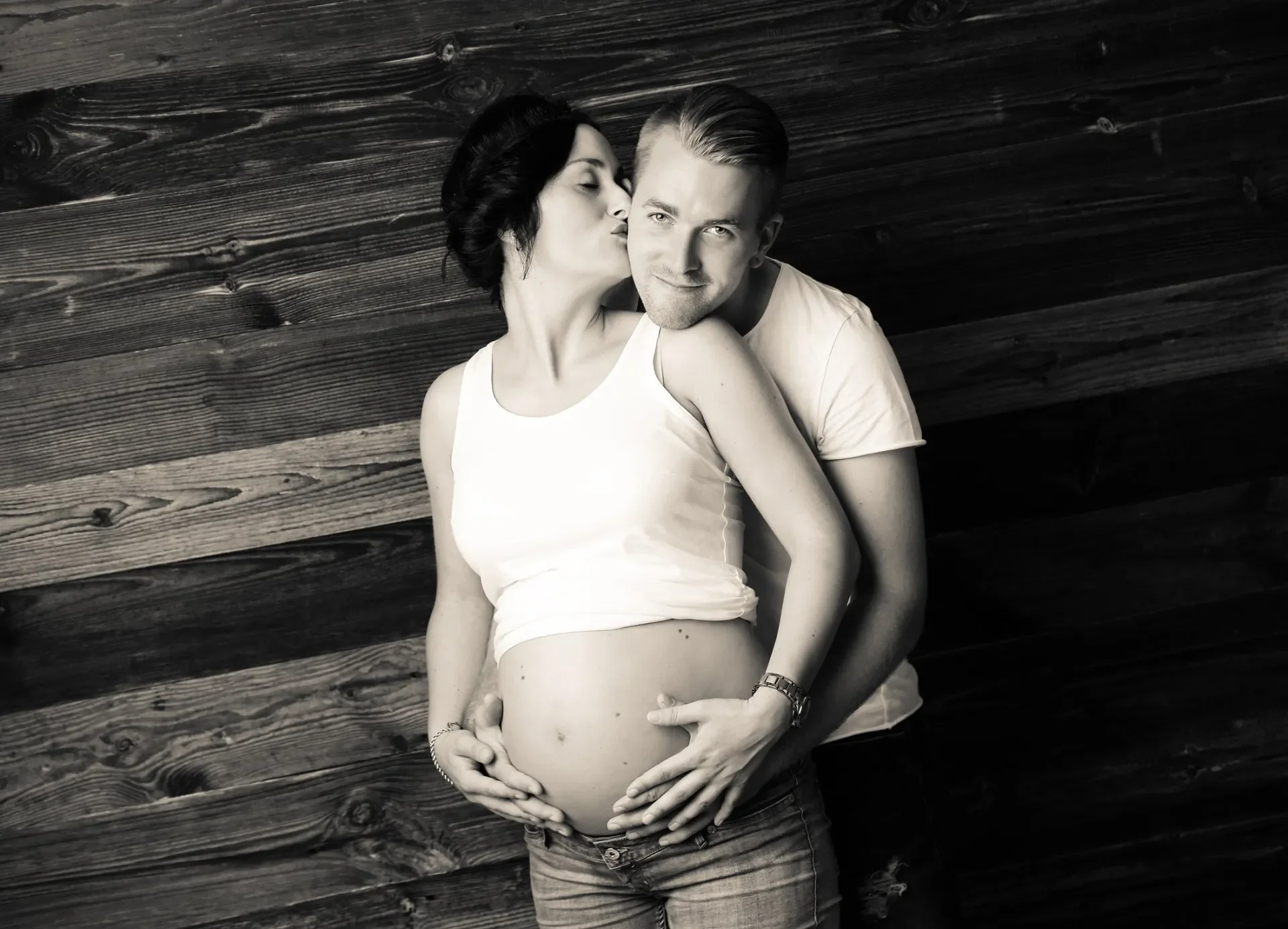 Babybauchfotoshooting Schwangere Frau mit Partner vor Holzwand, schwarz-weiße Fotografie, Frau umarmt ihren Bauch, Partner küsst sie auf die Wange.