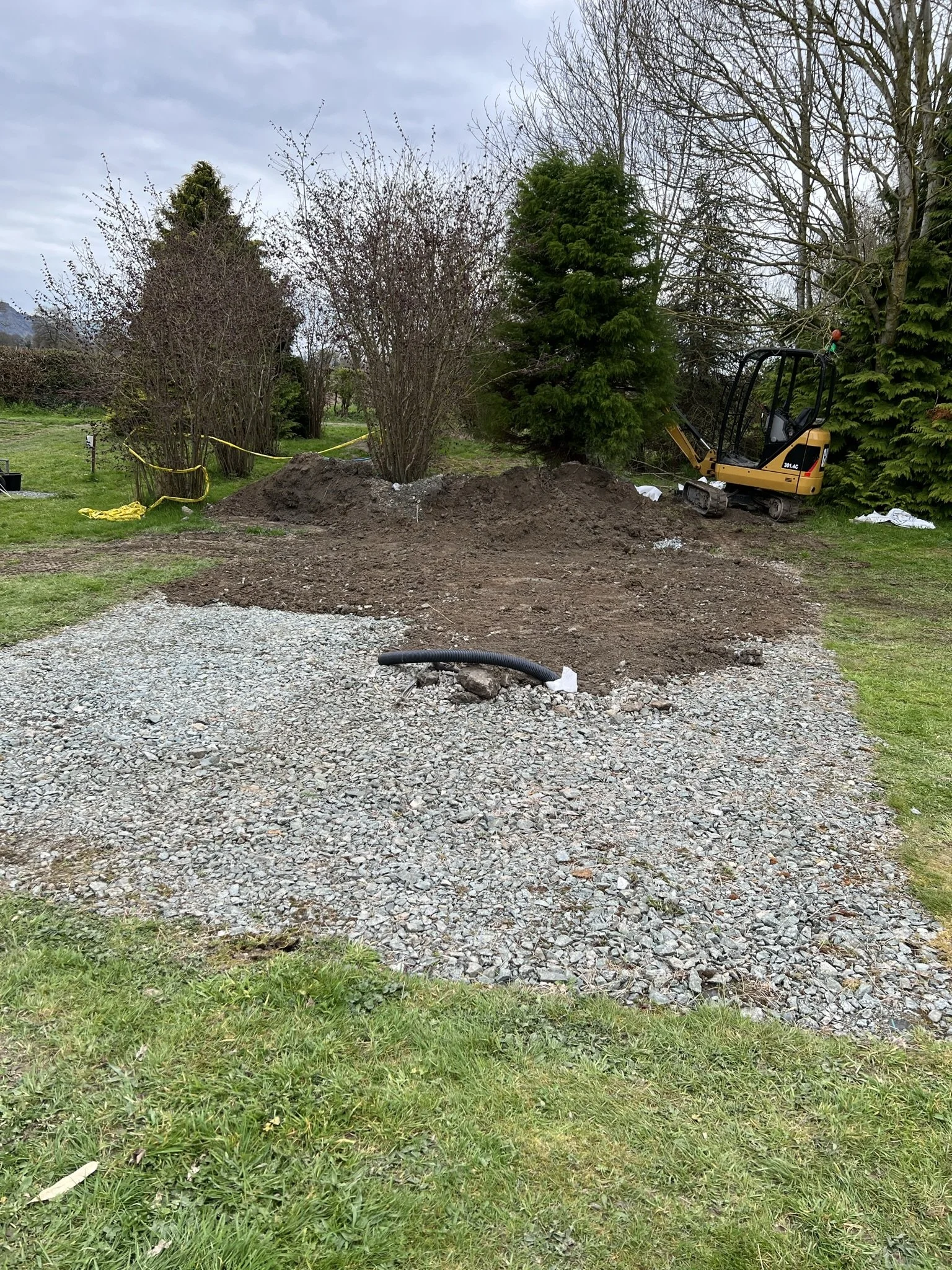 A small yellow excavator digging a hole in a backyard garden, surrounded by dirt piles and construction tape, with trees and bushes in the background.