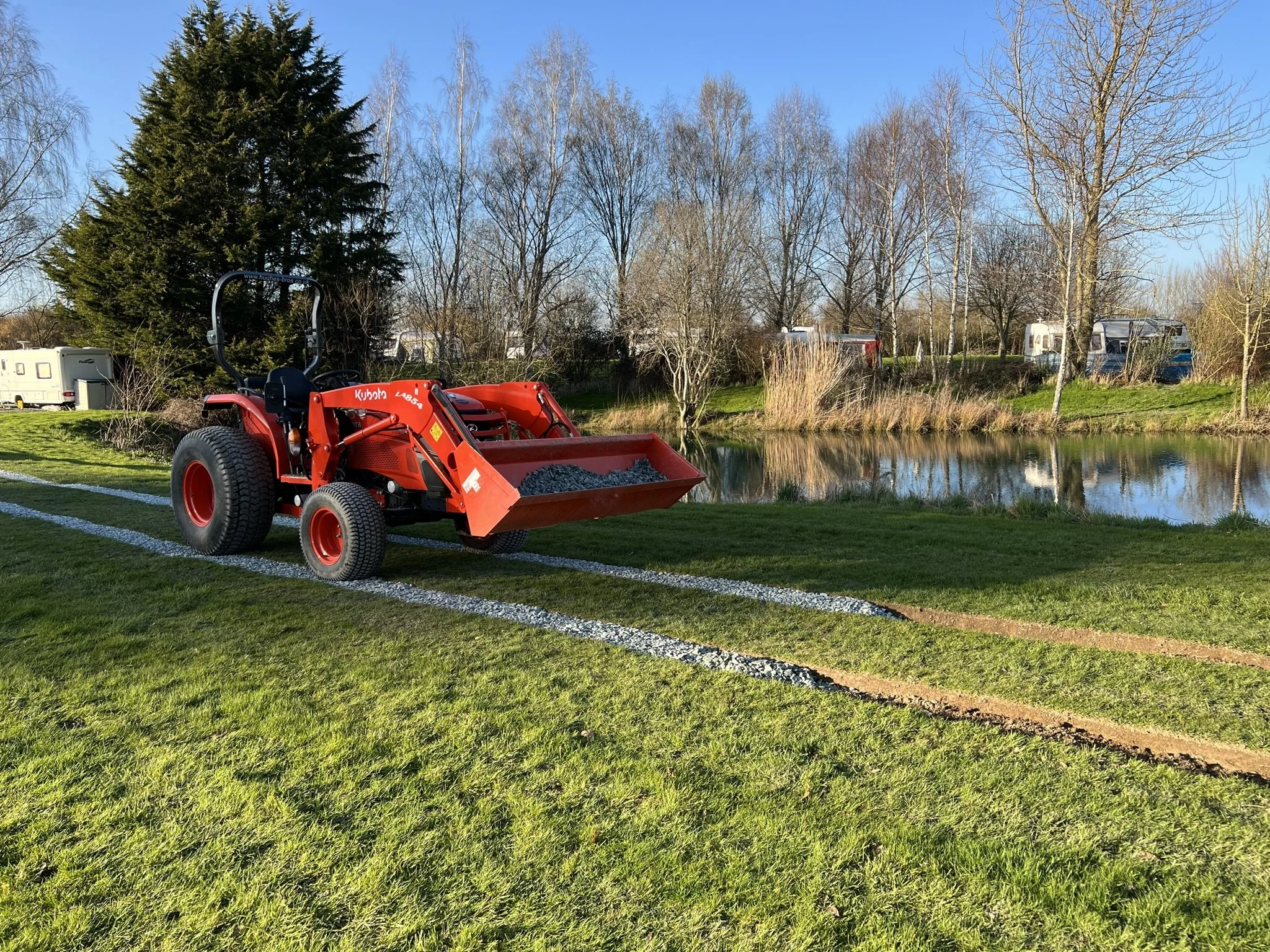 A red tractor with a front loader attachment filled with small rocks is parked on a grassy area next to a pond, with trees and caravans in the background under a clear blue sky.