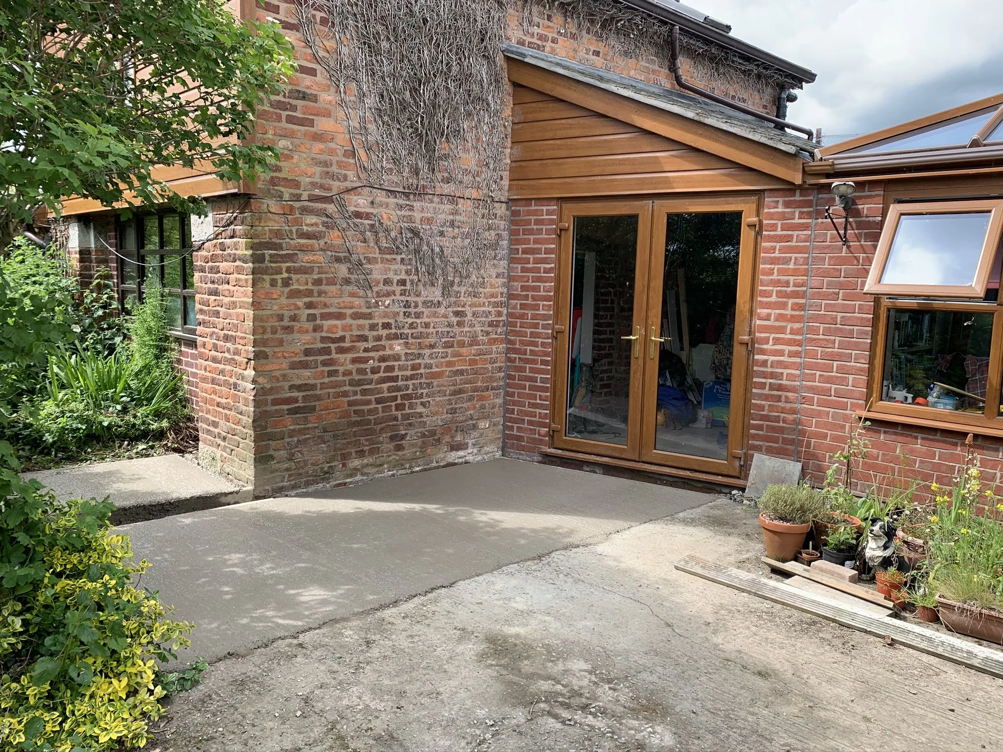 Backyard patio with new concrete slab, brick house with wooden and glass doors, windows, and garden pots with plants.