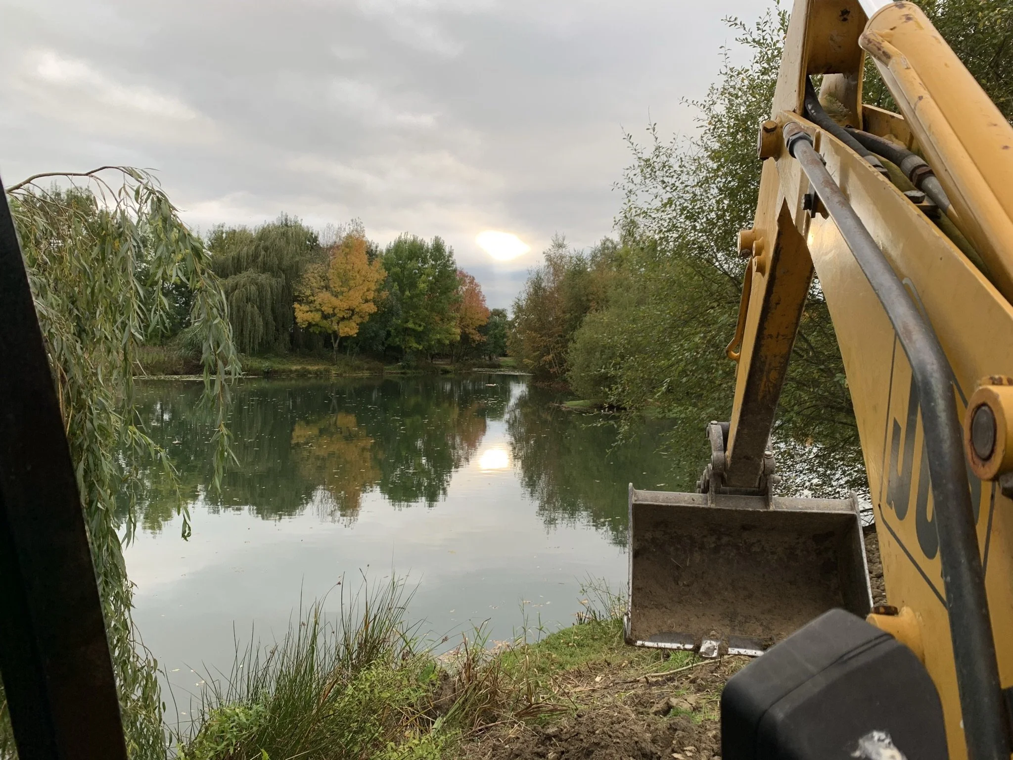 A yellow construction excavator near a body of water, surrounded by trees with autumn foliage, on a cloudy day.