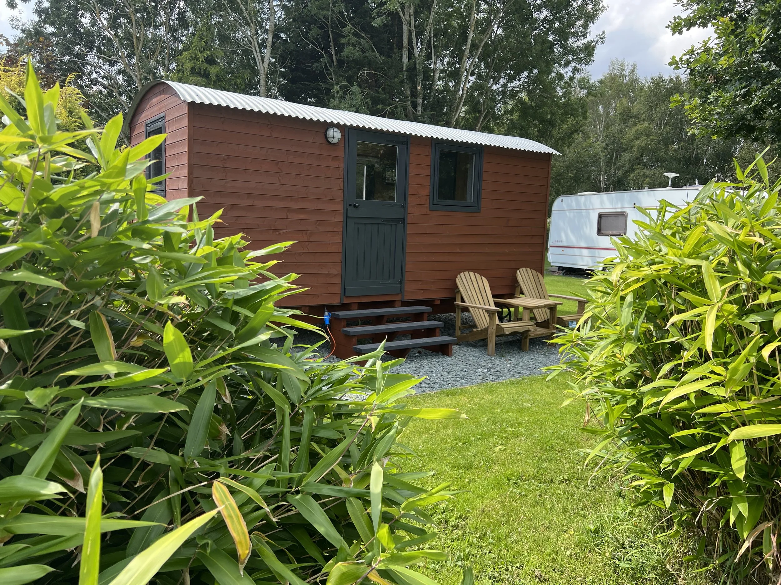 A small wooden cabin with a metal roof surrounded by green bushes and trees, with a white travel trailer in the background, in a lush outdoor setting.