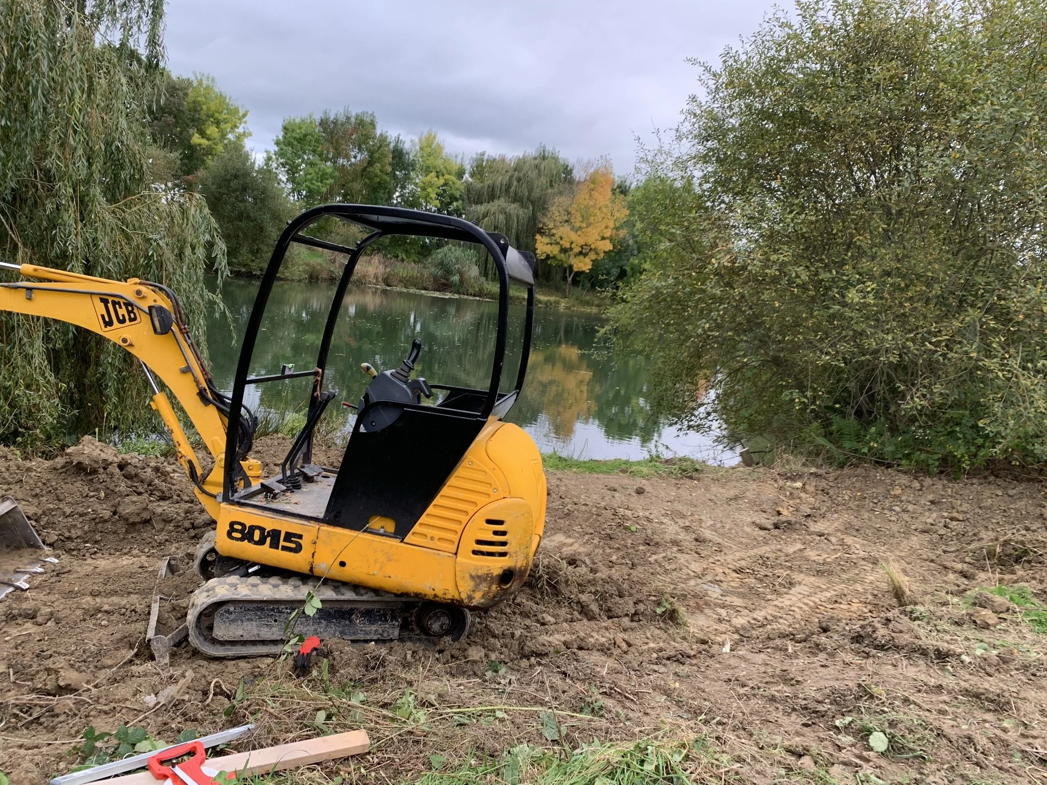 A yellow mini excavator on a dirt patch near a body of water, with trees and cloudy sky in the background
