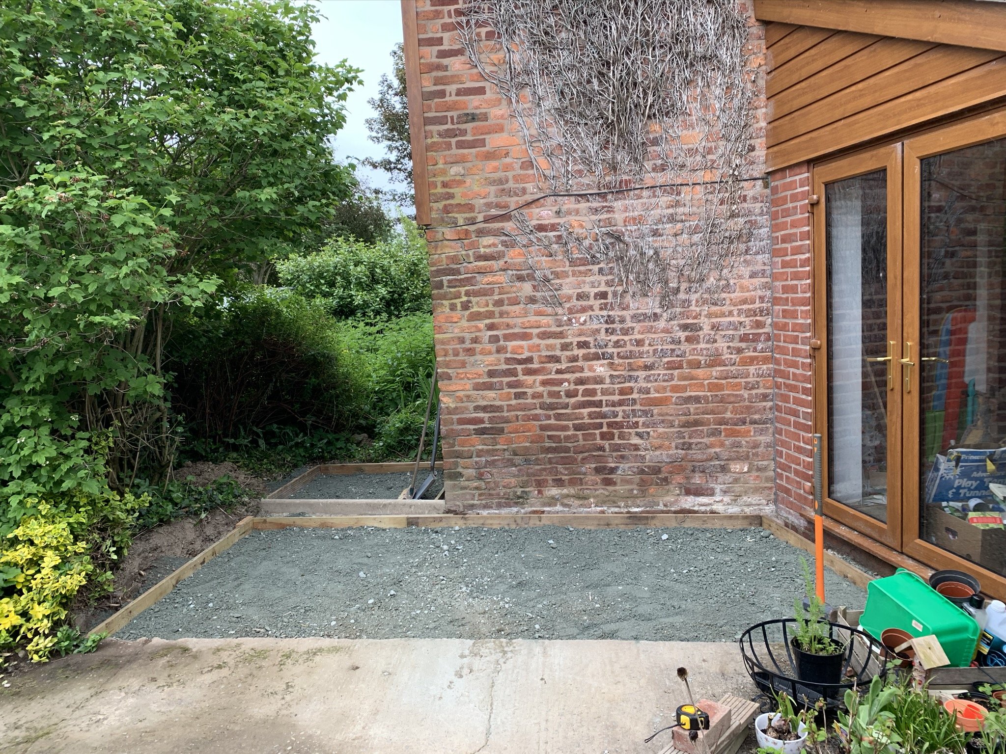 A backyard patio area under construction, with a gravel and dirt ground, wooden framing for a new deck or patio space, surrounded by lush green bushes and trees. There is a brick house wall with a vine growing on it and a sliding glass door leading inside.