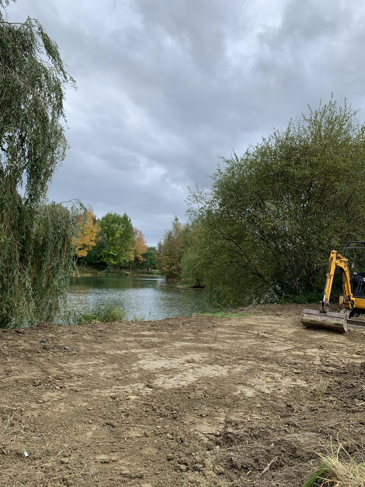 A small excavator parked on a dirt path by a river, with trees and overcast sky in the background.
