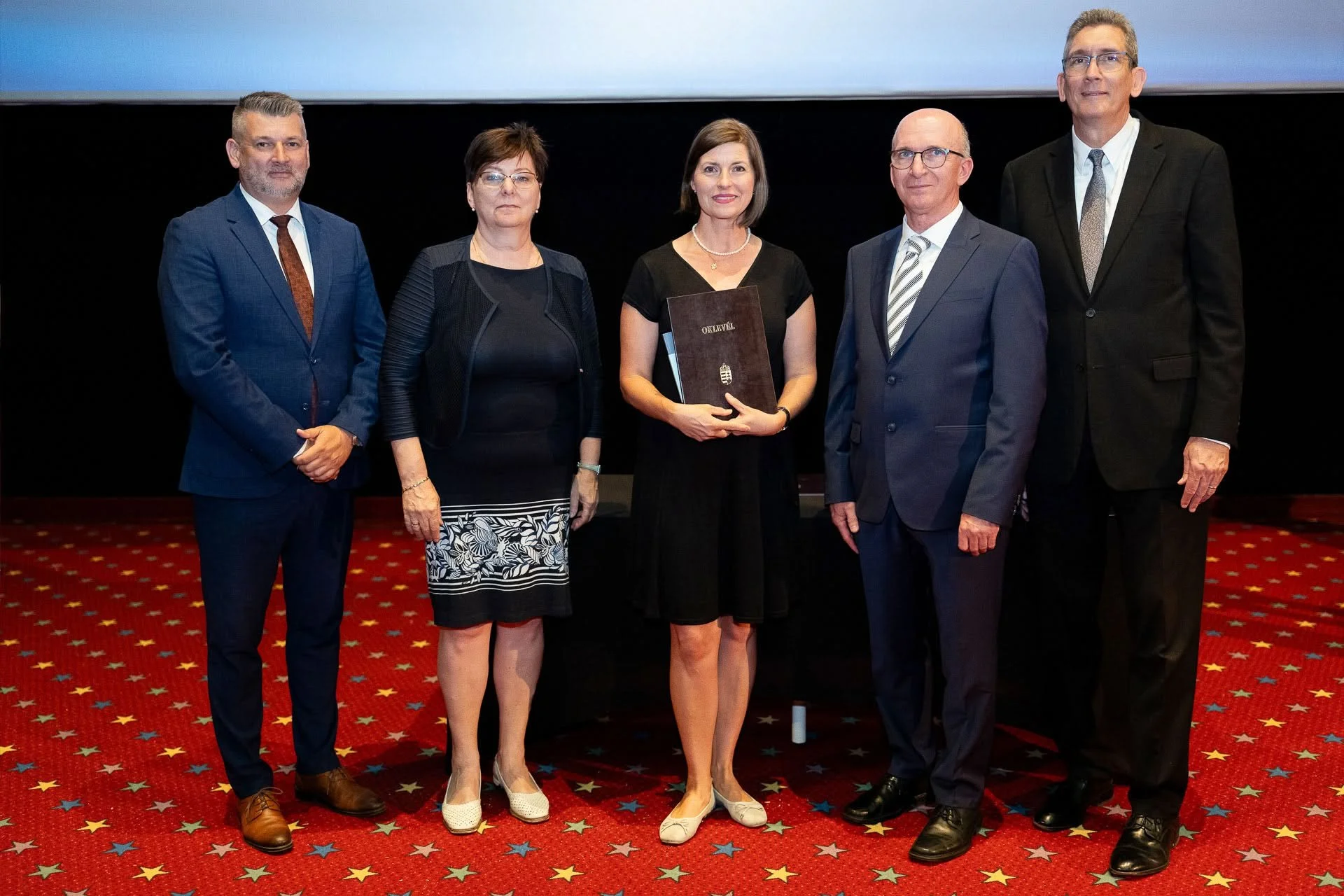 Nagy Erika PCC Coachery, a mentor coaching és coach szupervizor diplomájával a kezében, áll egy teremben, piros szőnyegen, körülötte a tanárai és a dékán, a diplomaosztó ünnepségen.