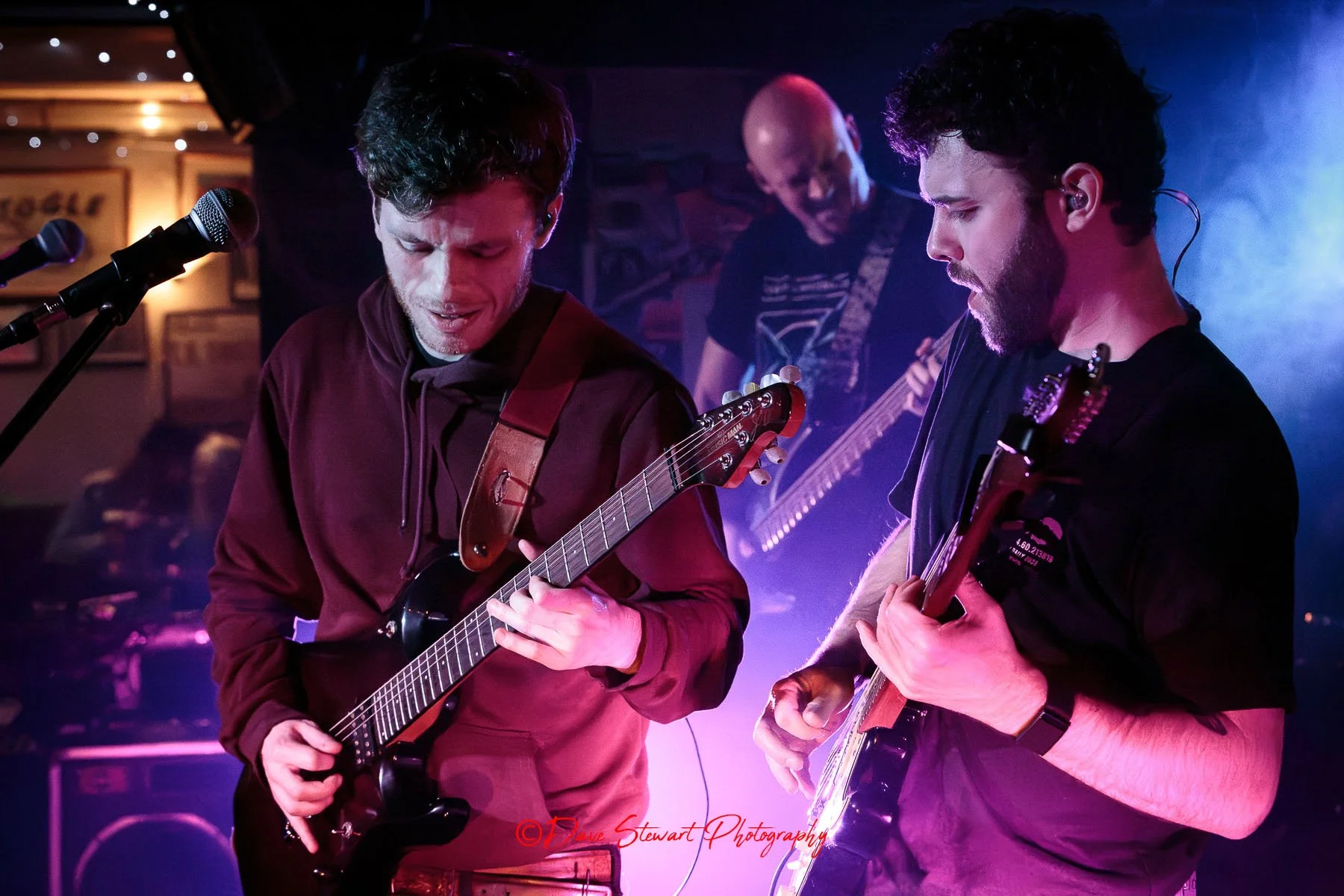 Two male musicians playing guitars on stage during a live performance, with a third musician in the background. The scene is lit with colorful stage lights, creating a vibrant atmosphere.