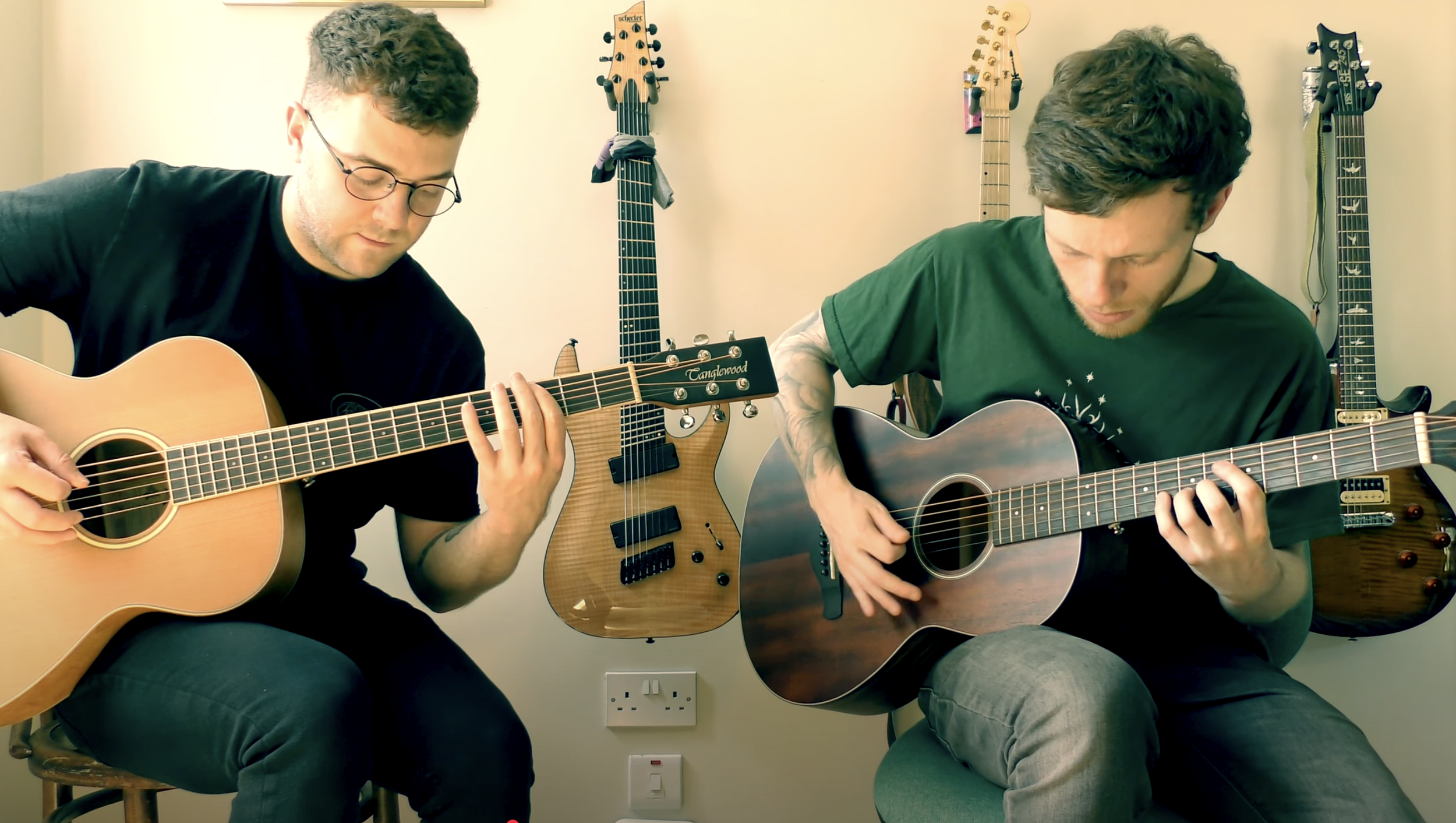 Two young men sitting side by side playing acoustic guitars, surrounded by multiple electric guitars hanging on the wall behind them.