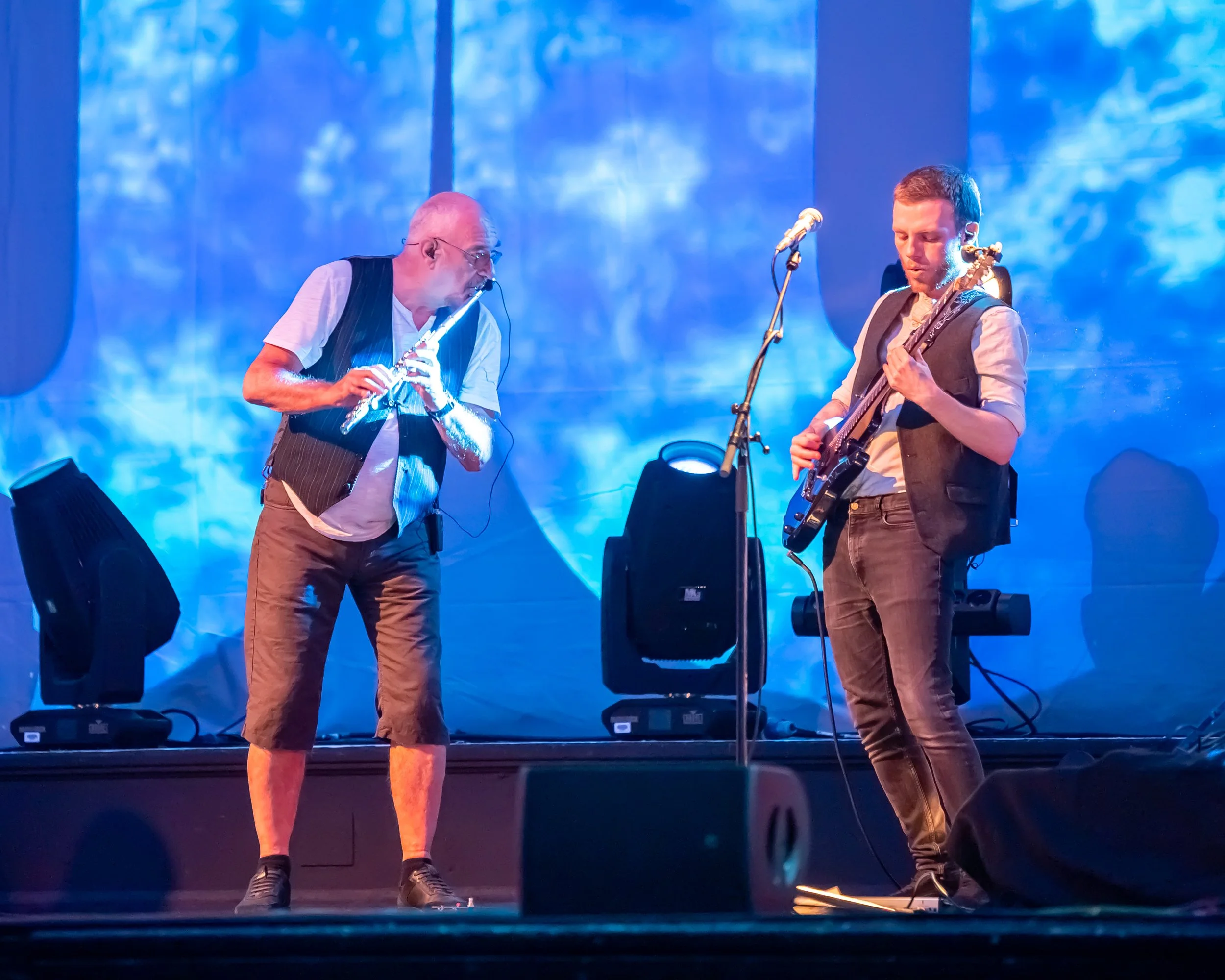 Two musicians performing on stage with blue lighting and a large backdrop. One is an older man playing a flute, wearing glasses, a white shirt, a black vest, and brown shorts. The other is a younger man playing an electric guitar, wearing a vest, a light-colored shirt, and brown pants.