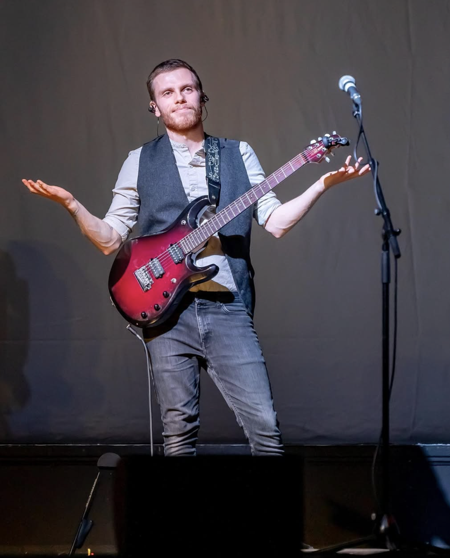 A man with brown hair and beard standing on stage with dark grey background, holding a red electric guitar, wearing a light grey shirt with rolled sleeves, a dark grey vest, and grey jeans, gesturing with shrugging shoulders, facing a microphone.
