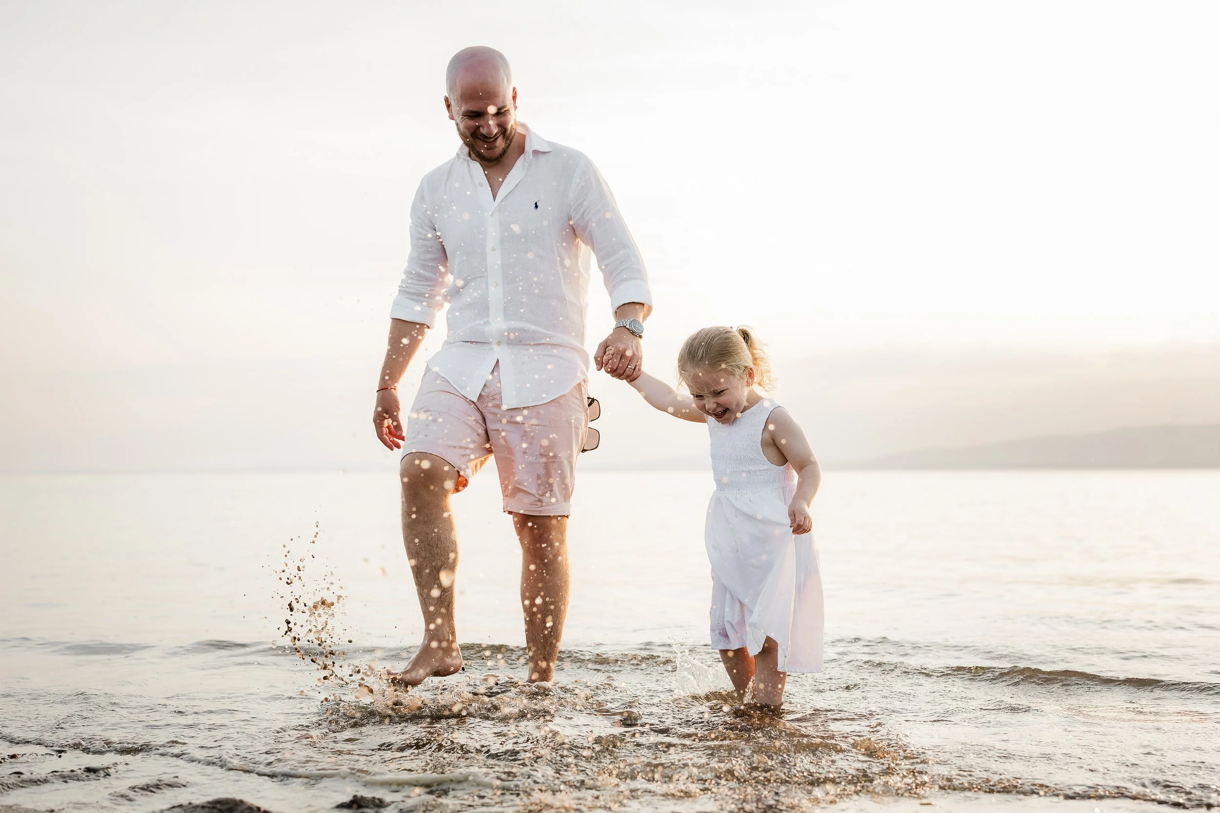 Dad and daughter holding hands and walking in shallow water at the beach during sunset, splashing water as they walk.