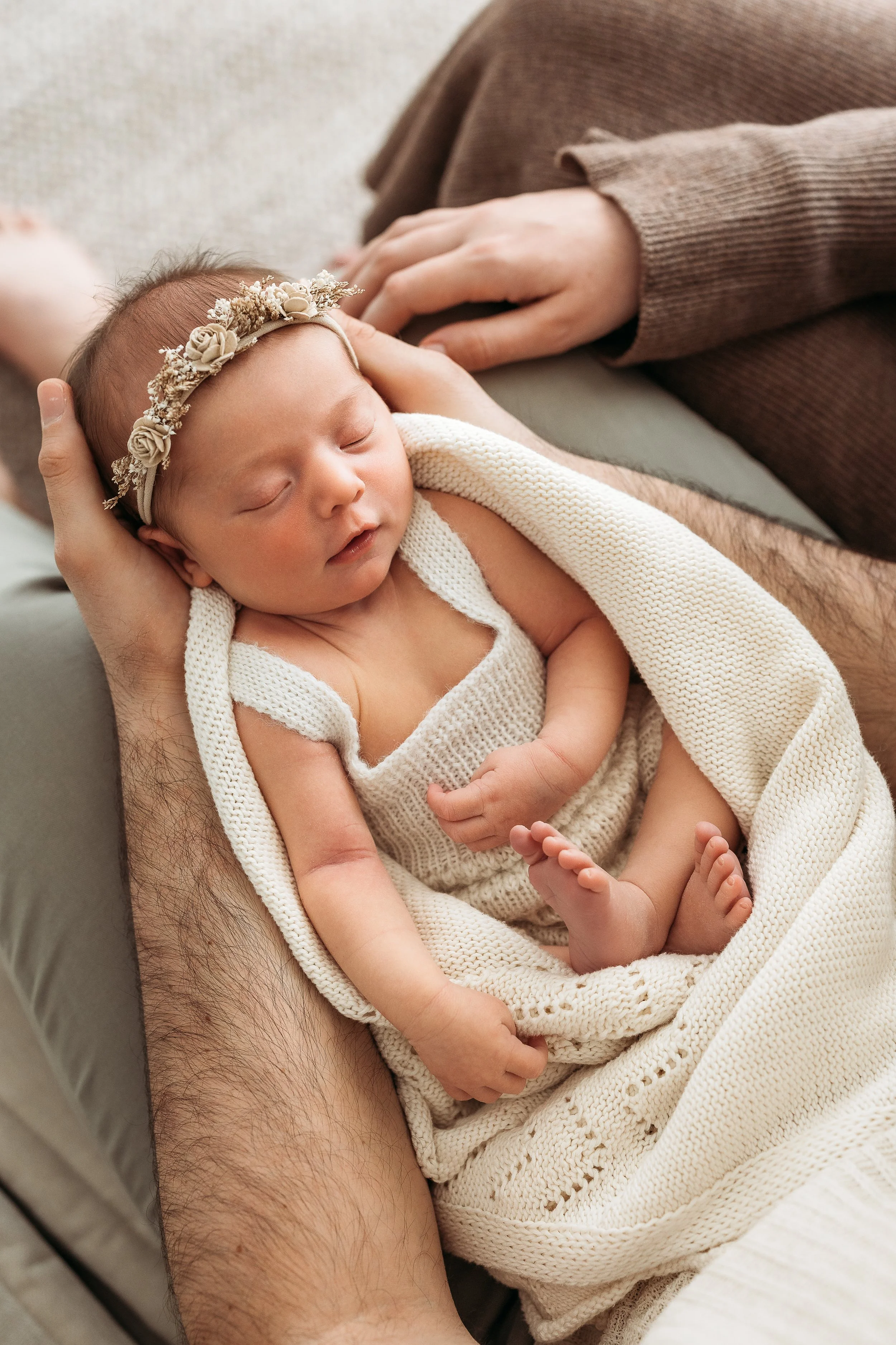 Sleeping newborn baby wrapped in a cream knitted blanket, cradled in a parent's arms during a natural newborn session.