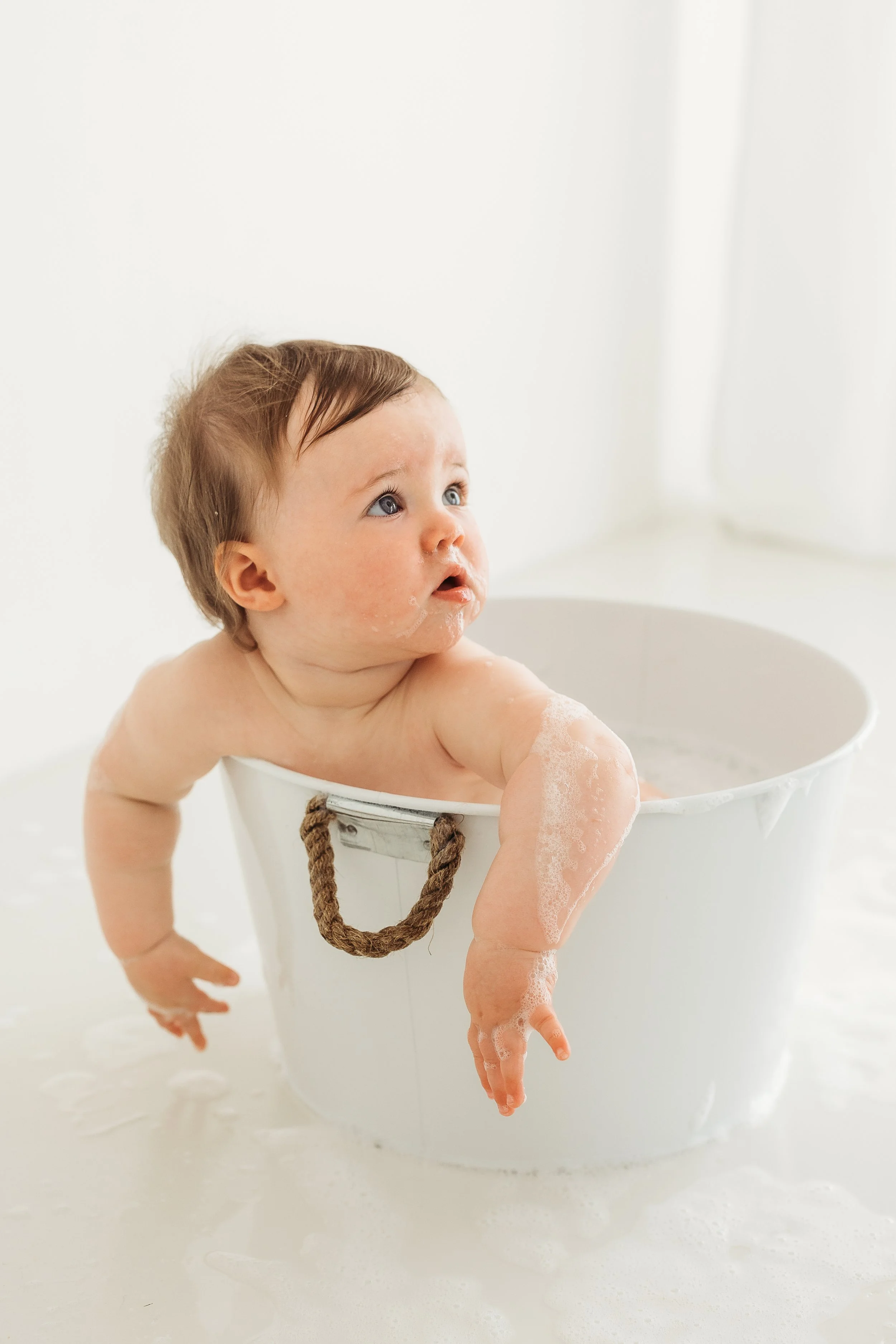 One year old child enjoying a bubbly bath in a white tub during a relaxed first birthday photography session