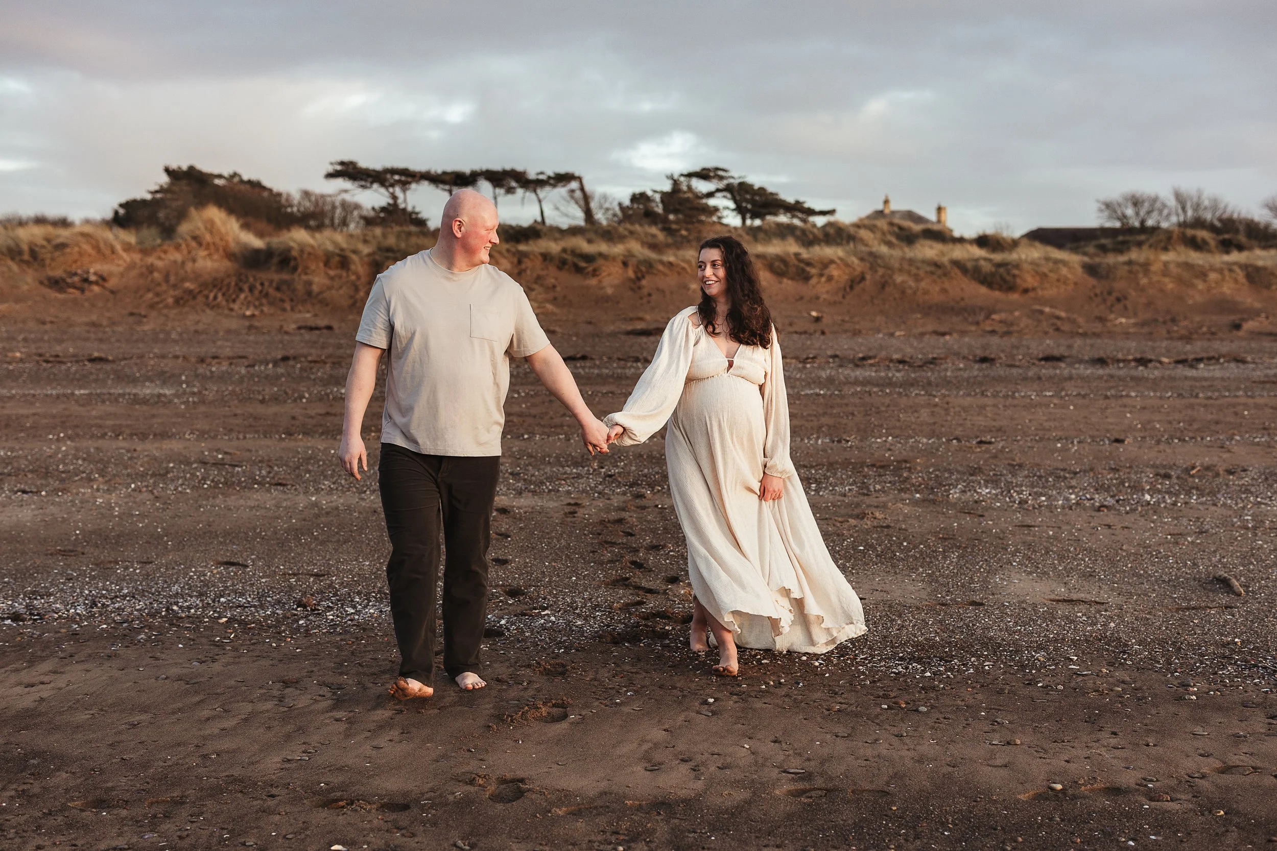 Expecting parents walking hand in hand along the beach during a relaxed outdoor maternity photography session.