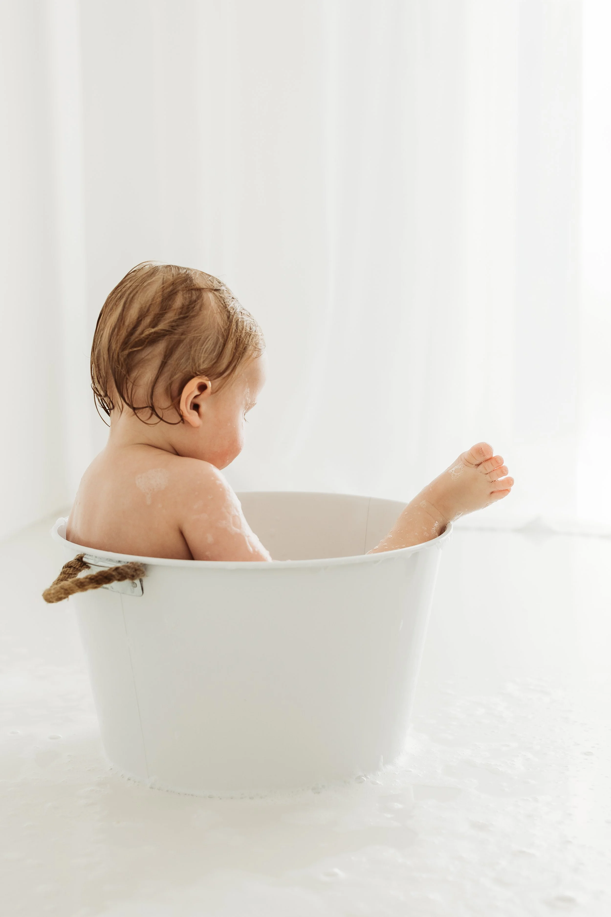 One year old child enjoying a bubbly bath in a white tub during a playful first birthday photography session