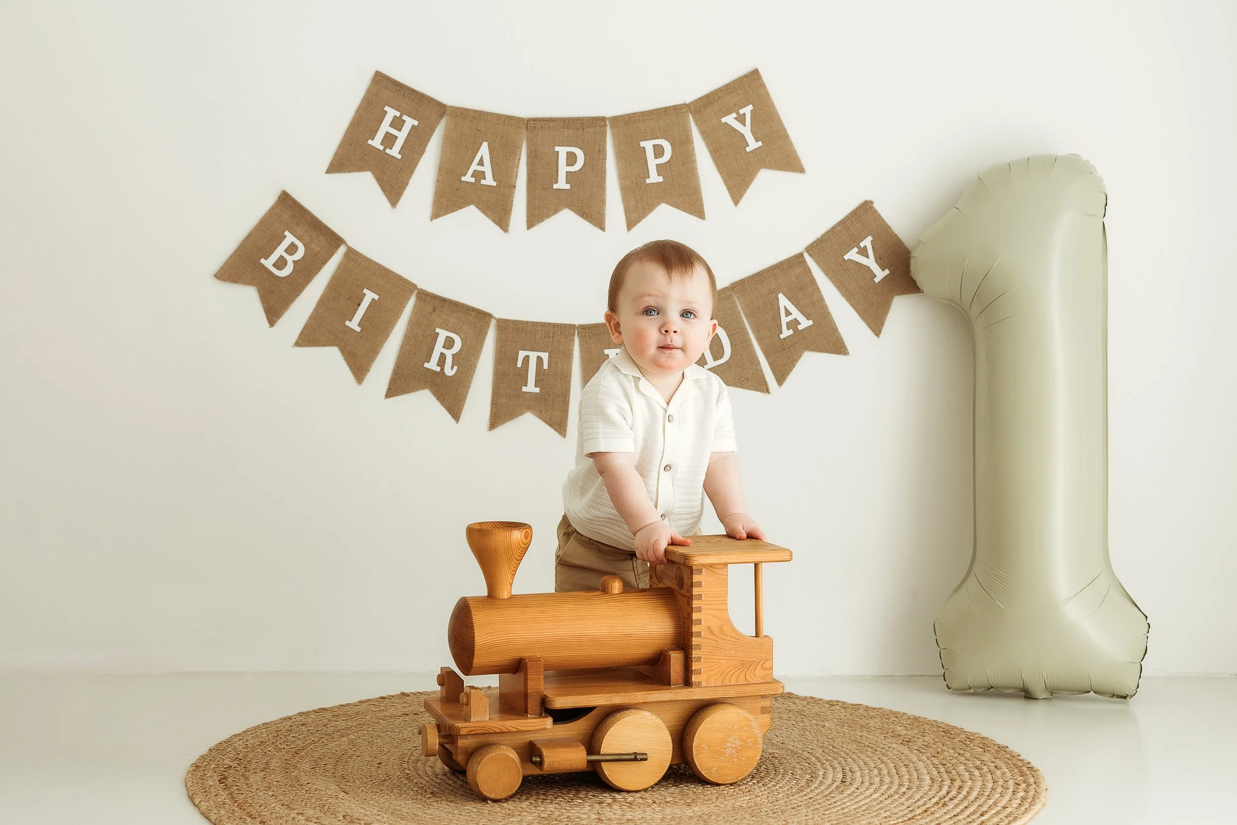One year old boy standing on a wooden train toy during a first birthday photography session with simple birthday decorations