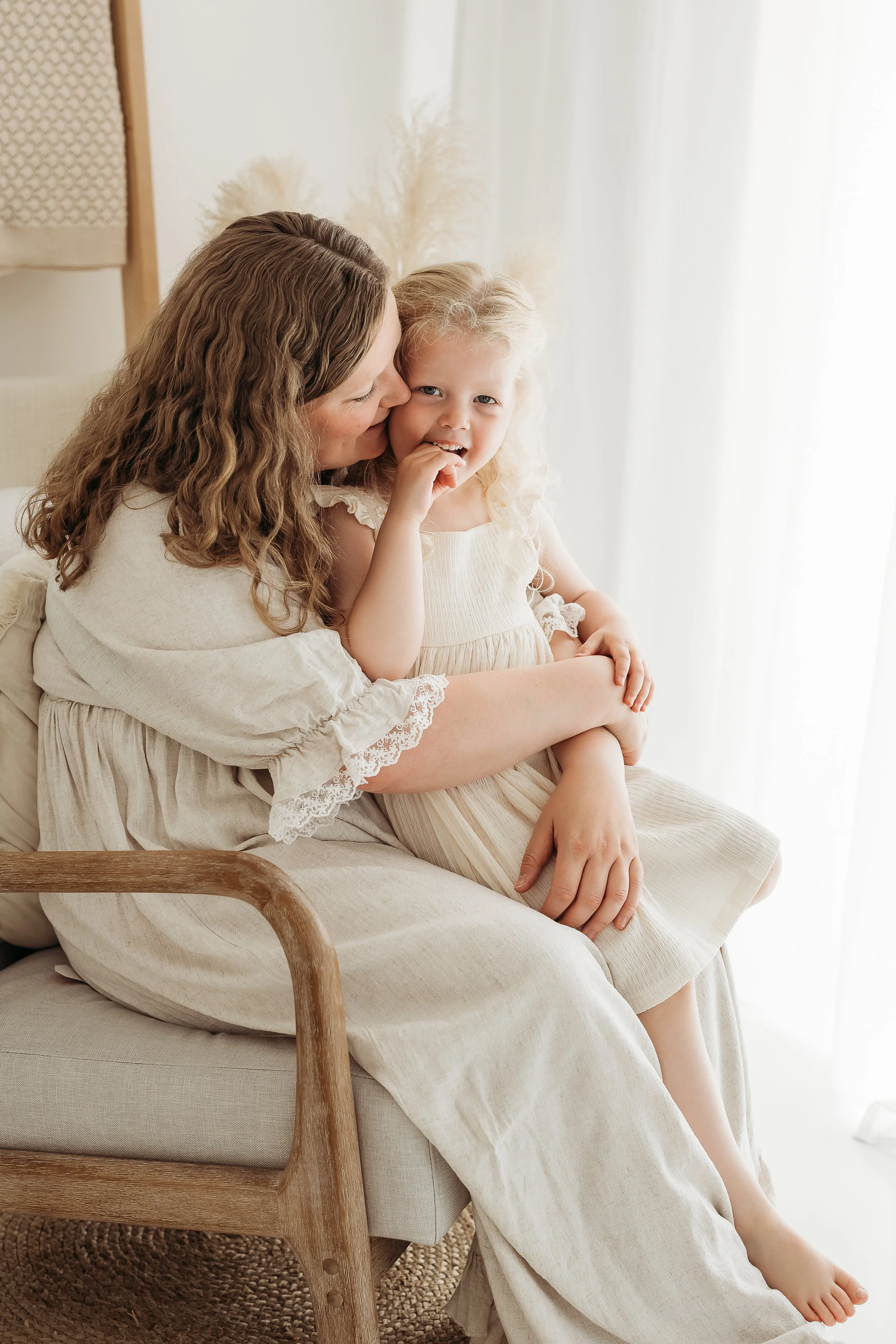 Mum cuddling her young daughter on her lap, both smiling together by a window.