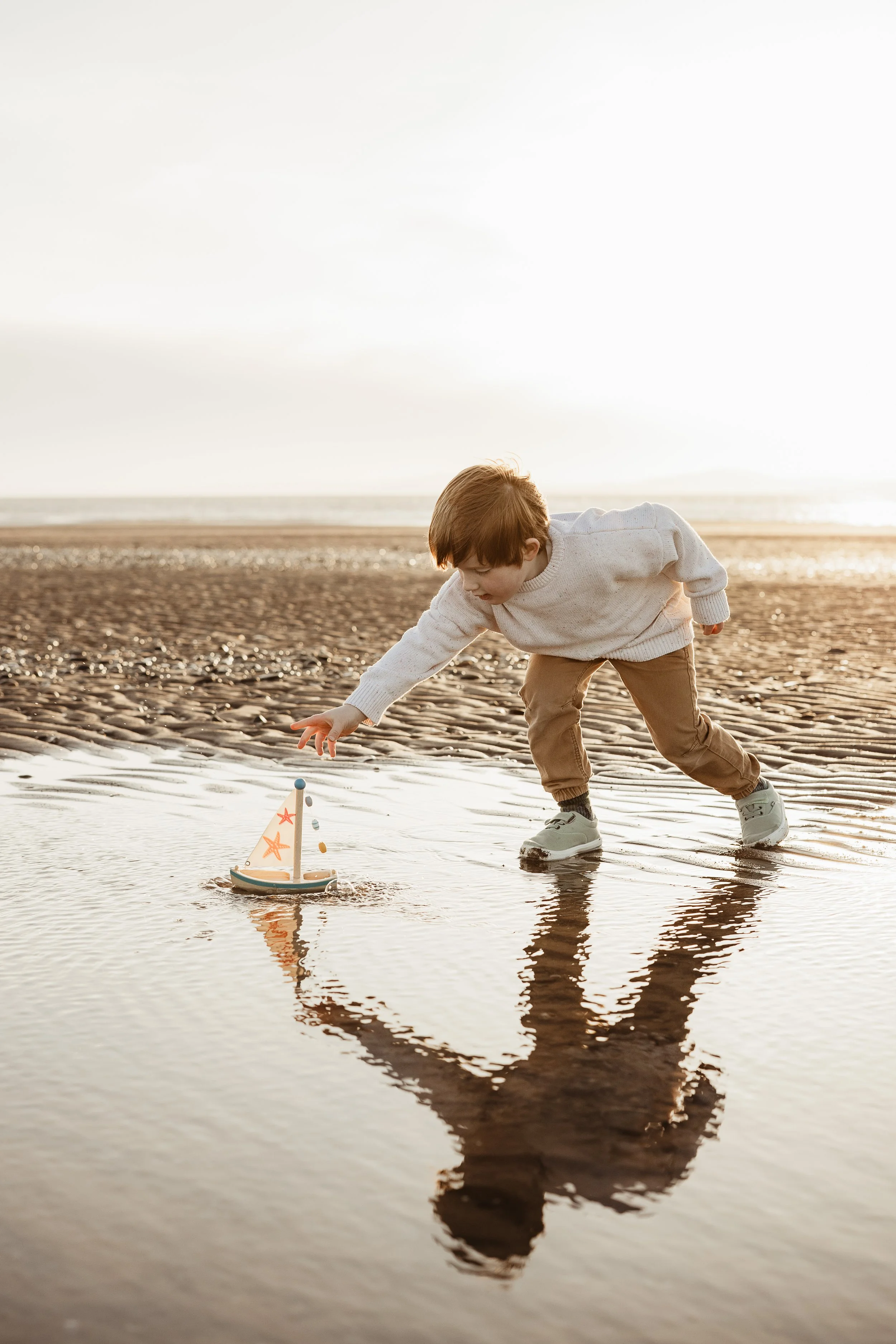 Young boy playing with a toy sailboat in the shallow water on Silloth West Beach at sunset.