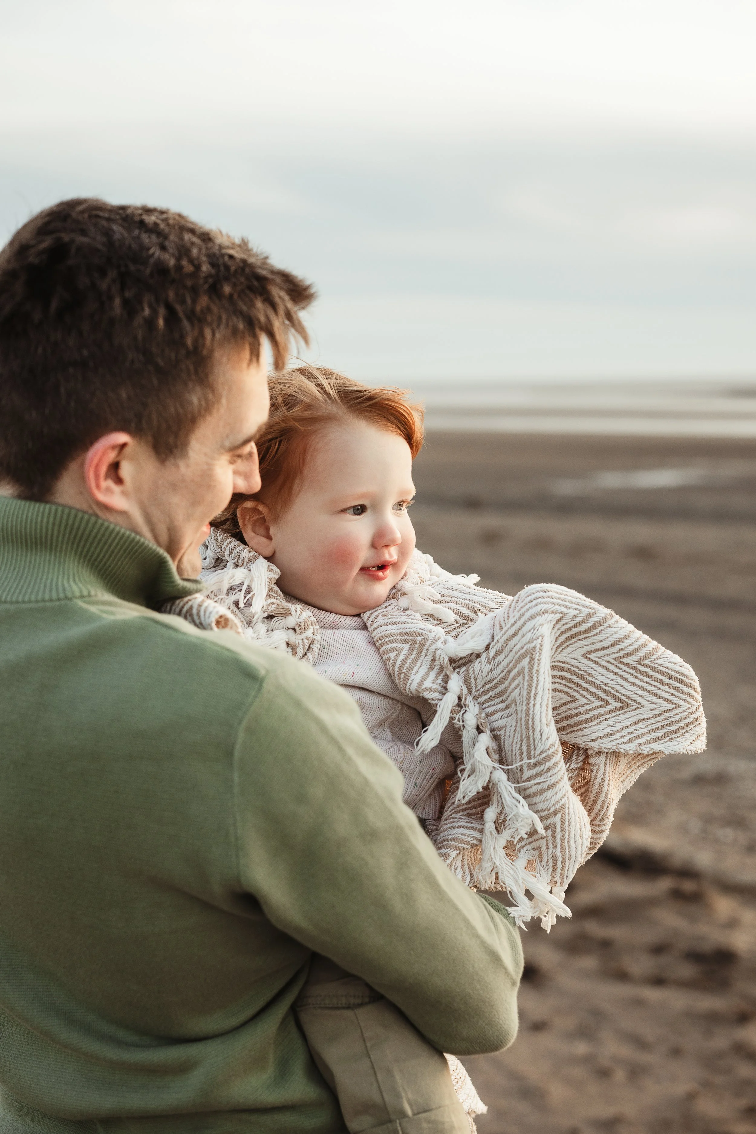 A father and son sharing a quiet moment together on the beach, looking out towards the sea during a relaxed family photography session.