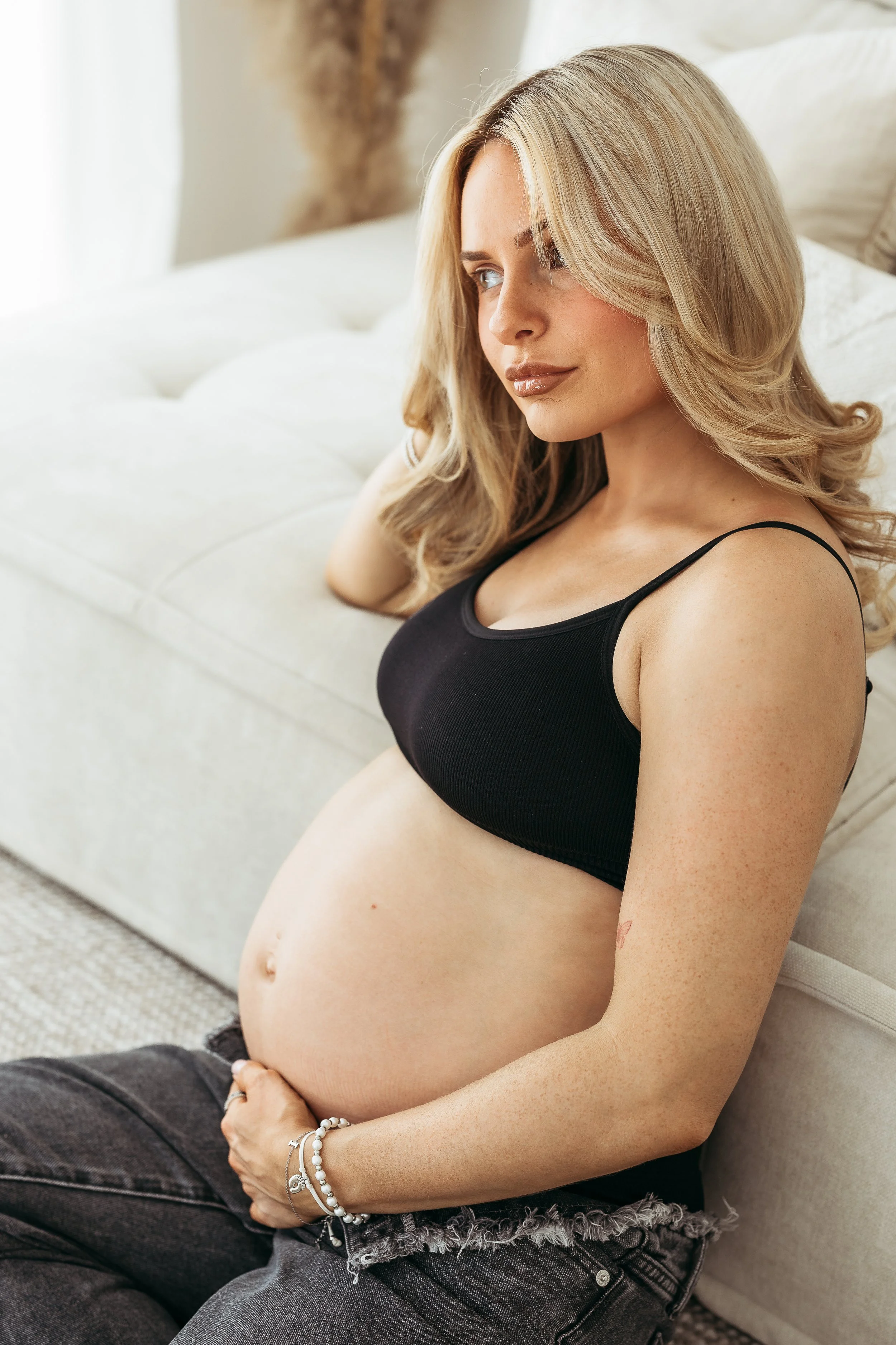 Pregnant woman sitting on the sofa and cradling her bump during a natural studio maternity session.