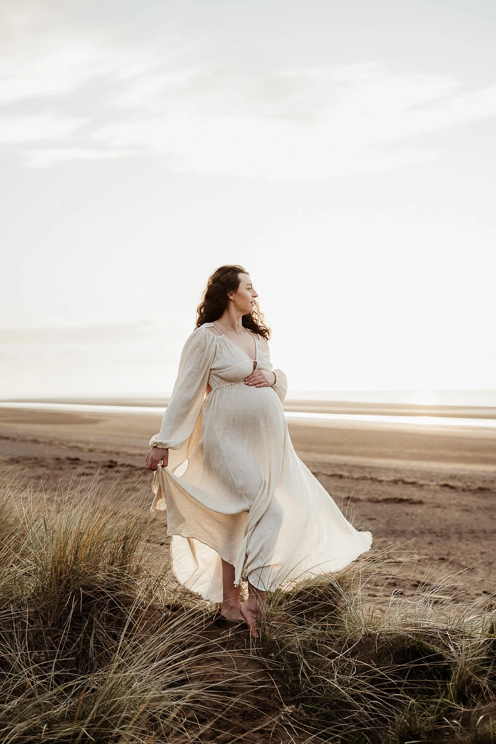 A mother-to-be walking barefoot through the dunes during a natural outdoor maternity photography session by the coast.