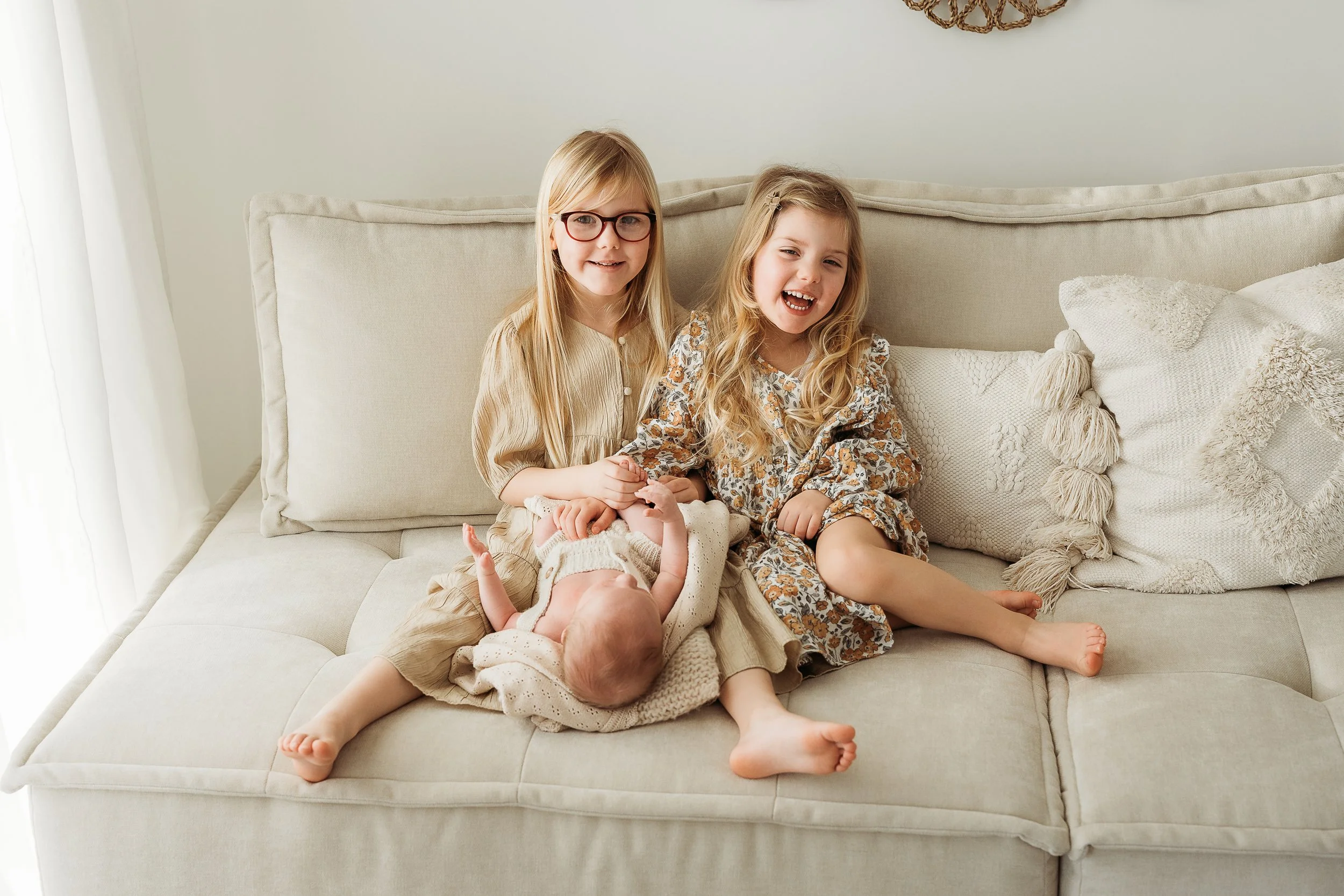 Two sisters laughing and cuddling their new baby sibling during a relaxed newborn session.