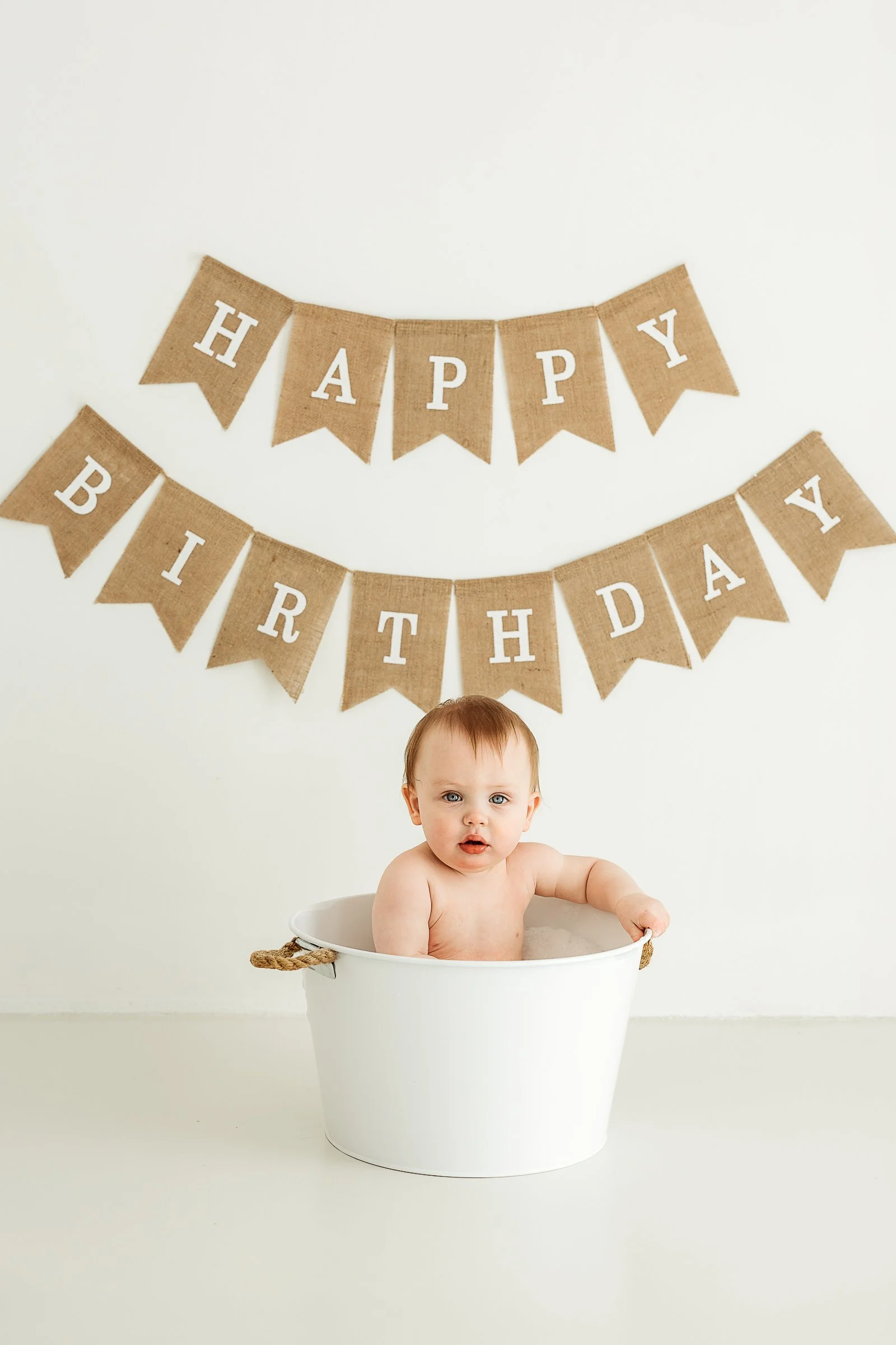 Baby sitting in a white bucket with a birthday banner during a playful first birthday photography session