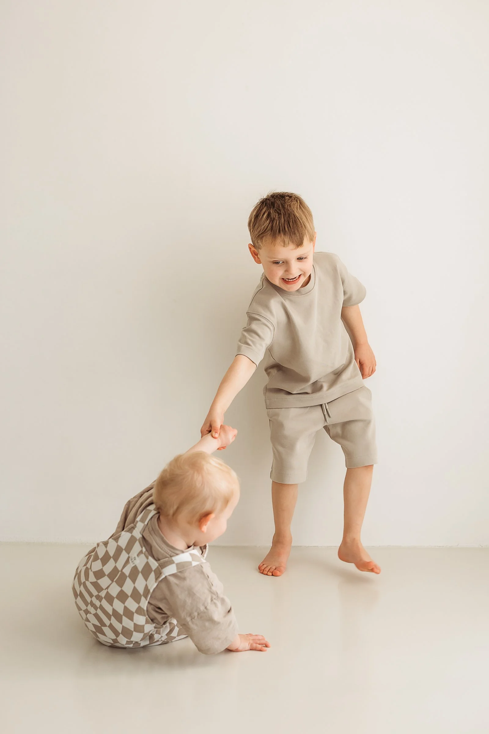 Two young boys, one standing and smiling, the other sitting on the floor, holding hands in a minimalistic room with a plain white wall.