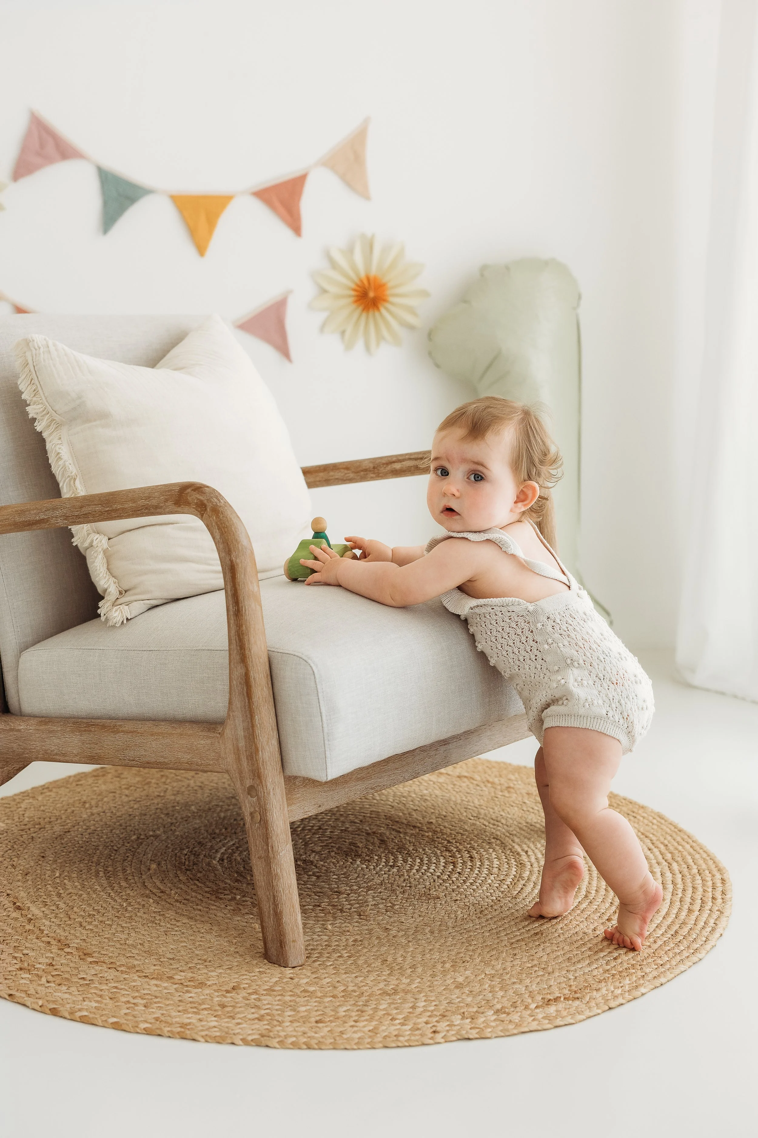 One year old baby standing beside a chair in a bright natural light studio during a first birthday photography session