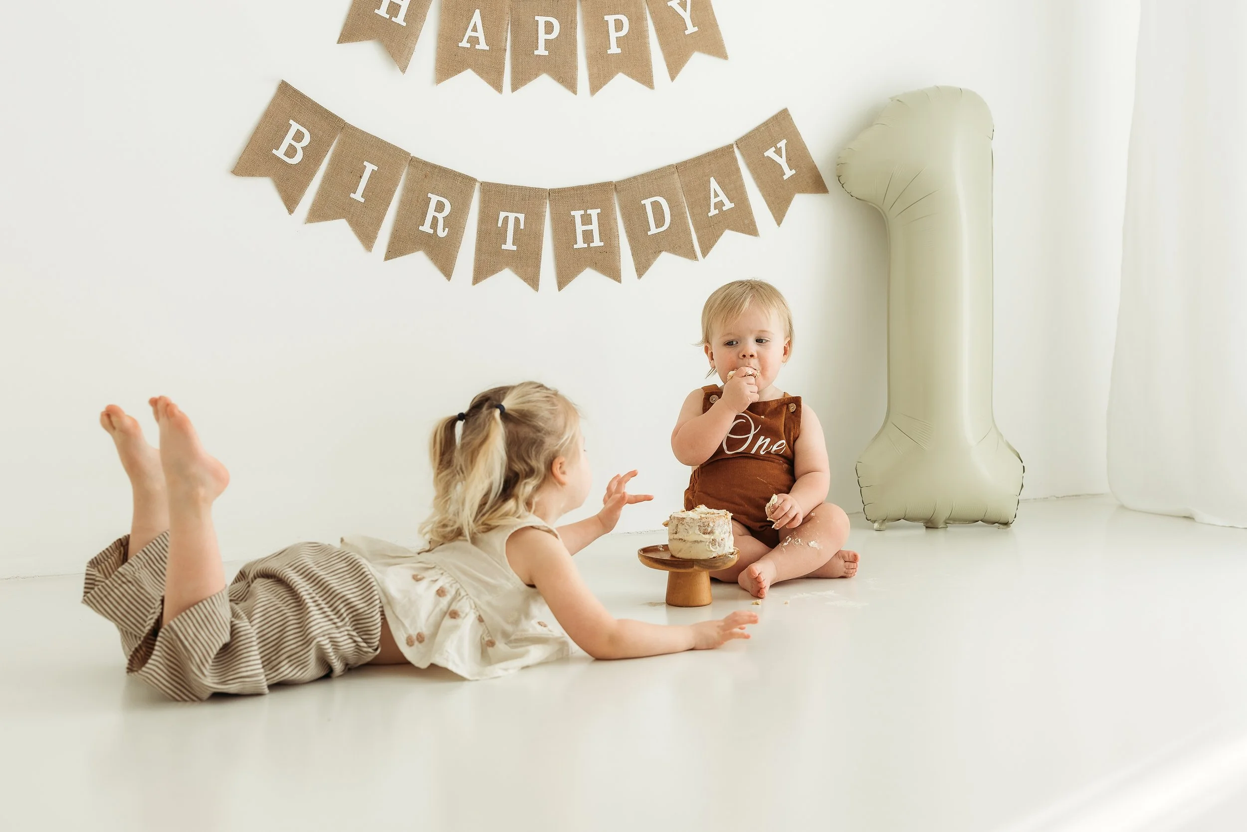 Siblings celebrating a first birthday with cake, a number one balloon and birthday decorations in a natural light studio